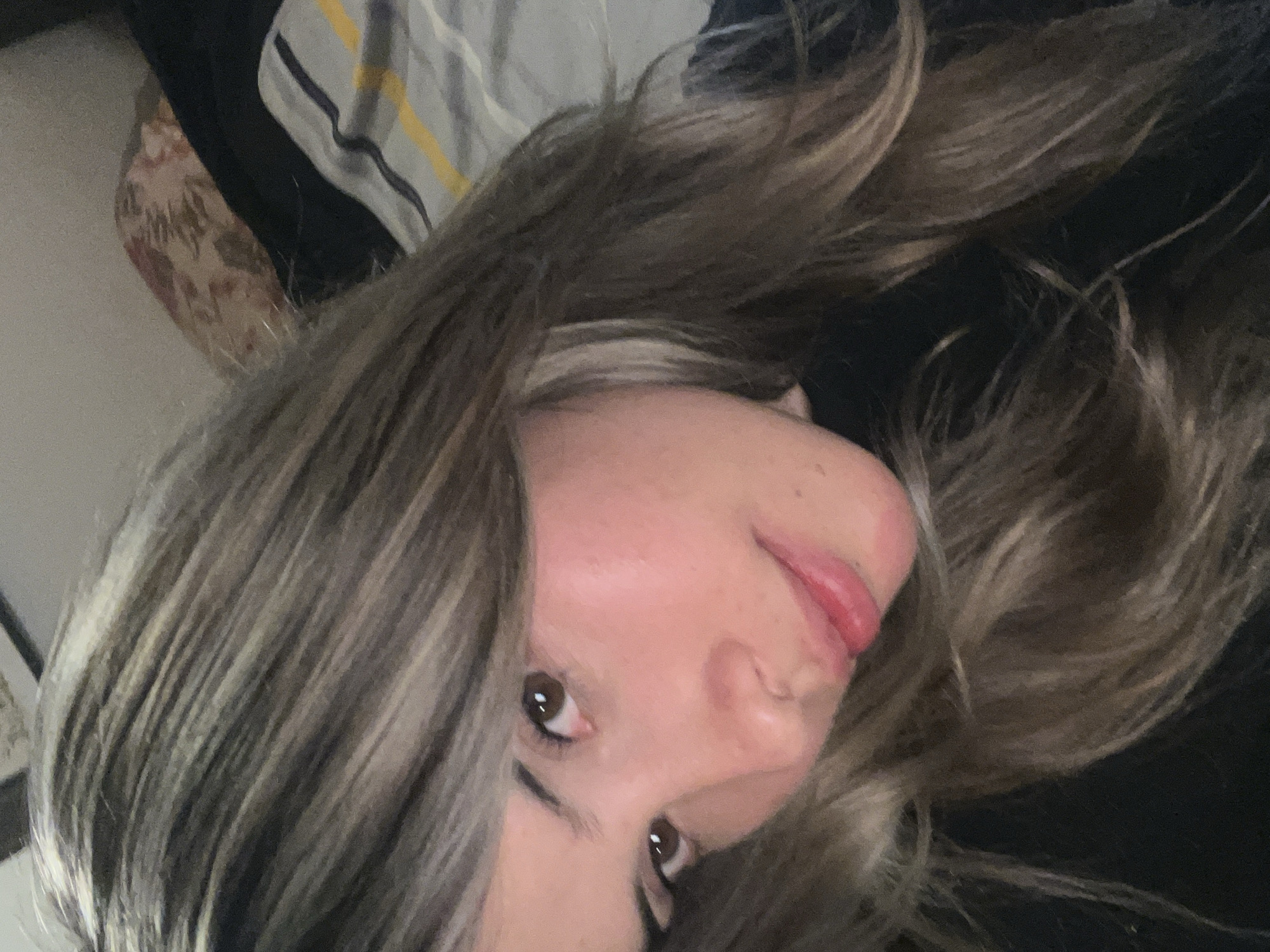 A close-up selfie shows a woman with long, brown hair with blonde highlights, looking at the camera. She is wearing a black shirt. Her face is illuminated by soft light, highlighting her brown eyes and pink lips. The background is blurred, but appears to be a bed with striped sheets and a patterned pillow. There is no indication of the city of Sumalao, Argentina in the image.