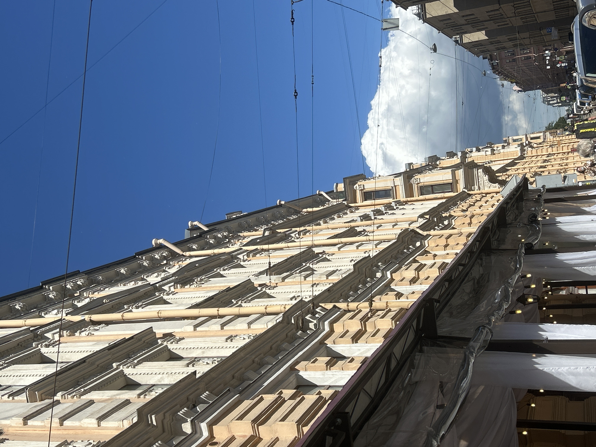 The image depicts a street scene in a city, likely on a sunny day with a bright blue sky and scattered clouds. On the left side of the frame, a tall, ornate building with beige and white detailing stretches along the street. The building features many windows and decorative architectural elements. In front of this building, there is an awning covered with clear plastic sheeting, with white fabric drapes hanging behind it. Several string lights are visible, illuminating the space beneath the awning.
The street itself is lined with buildings, and power lines crisscross the sky above. In the distance, more buildings can be seen, receding into the background. Several cars are parked along the street, and a few people are visible walking in the distance, appearing as small figures in the urban landscape. One person in the foreground is partially obscured, wearing a mask and holding a phone. The overall impression is of a well-established urban environment with a blend of historical architecture and modern street infrastructure.