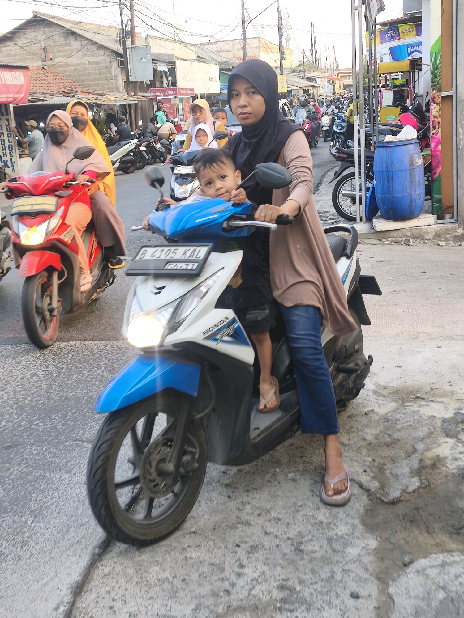 In Satriamekar, Indonesia, a woman wearing a dark hijab and a tan cardigan rides a blue and white Honda motorcycle. She is holding the handlebars with her right hand, and her left hand is placed on the back of a young child seated in front of her. The child, wearing a dark shirt and shorts, looks towards the camera. The woman's legs are clad in dark blue jeans, and she wears flip-flops. The motorcycle's license plate reads "B 4195 KAL". Another motorcycle, red in color, is visible to the left, carrying a woman in a yellow hijab and a child. The background shows a street scene with buildings, overhead wires, and other vehicles, suggesting moderate traffic. The ground is paved with asphalt and concrete, with some visible cracks and debris. The overall lighting indicates it is daytime, possibly late afternoon.