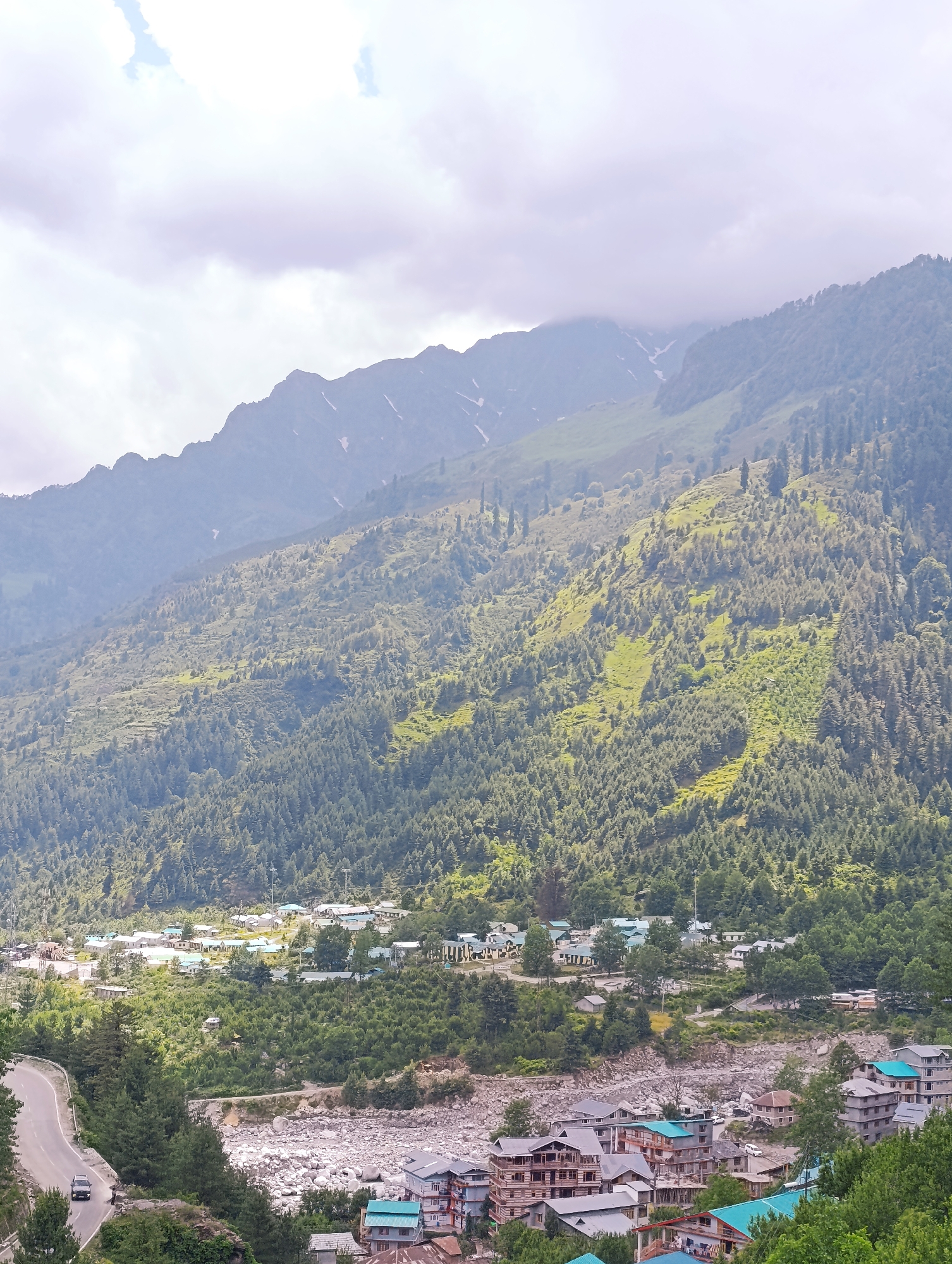 The image captures a panoramic view of Palchan, India, showcasing a settlement nestled amidst lush green mountains. The landscape is dominated by a winding road, a rocky riverbed, and a cluster of buildings, including residential structures and a few commercial establishments. Verdant pine forests carpet the slopes of the mountains, with patches of sunlight illuminating the greenery. The sky is partly cloudy, with a muted light suggesting an overcast day. A solitary vehicle is seen driving on the road, contributing to the serene atmosphere of the scene.