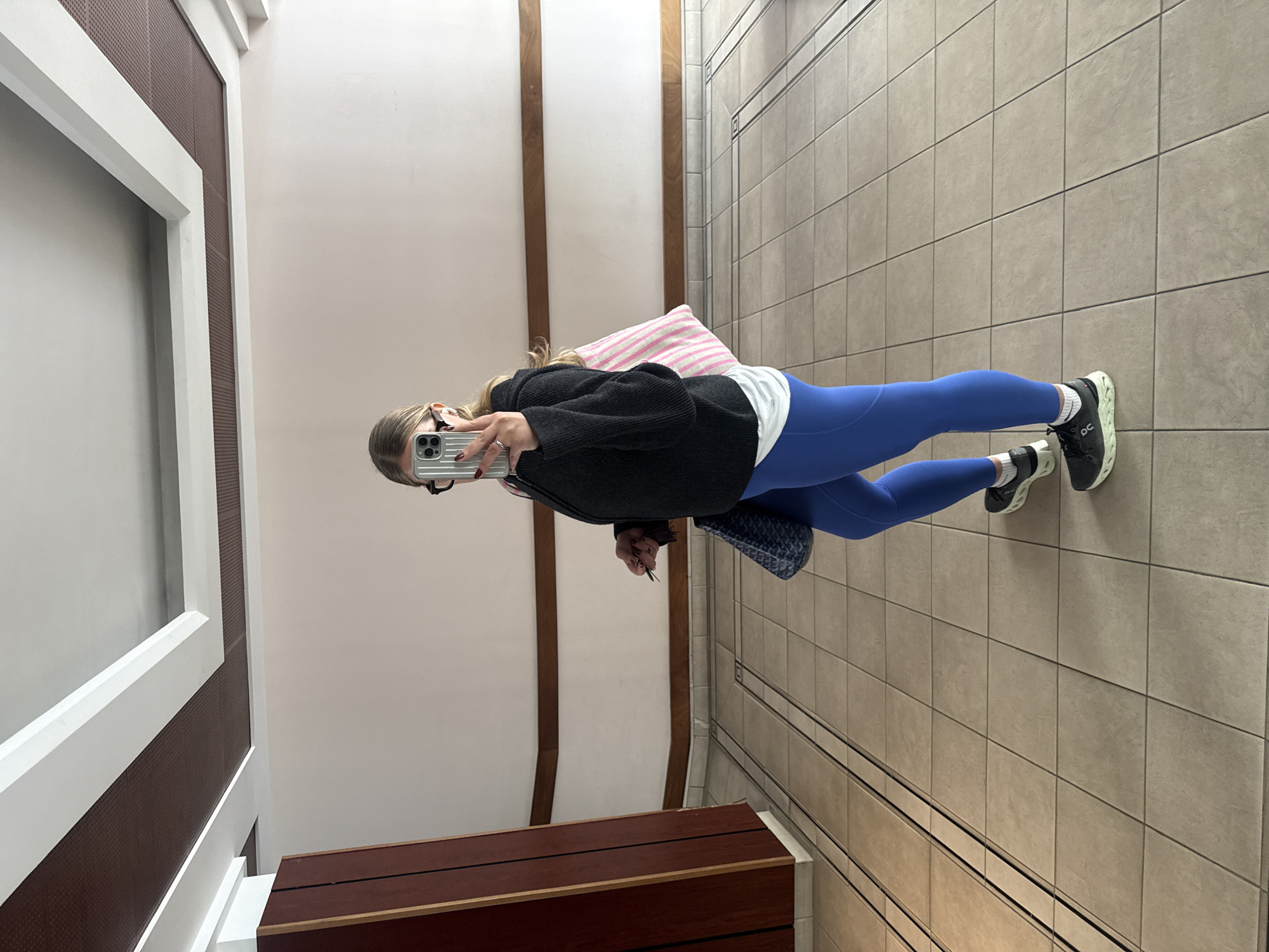 A person is standing in what appears to be an interior hallway of a building, taking a selfie. The person is wearing blue leggings, a white shirt, a dark-colored jacket, and sneakers. They are holding a smartphone up to take the picture. The environment includes tiled flooring and neutral-colored walls. There are wooden accents on the walls and a built-in seating area with a dark wooden top. The person has a shoulder bag and is holding keys. The location does not appear to be in Paris, France.