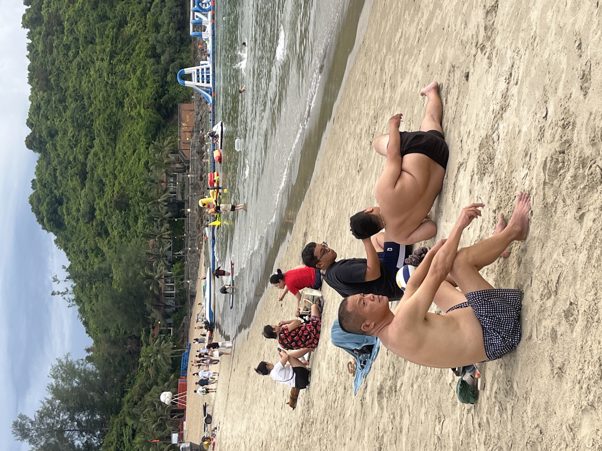 The media file shows a beach scene in Da Nang, Vietnam. Several people are relaxing on the sandy shore, some lying down and others sitting. The ocean is visible in the background, with gentle waves and a few people swimming or wading in the water. Further back, there's an inflatable water park with slides and other structures. The shore is lined with lush green trees and vegetation, suggesting a tropical environment. The sky is overcast, indicating a cloudy day. The people are dressed in casual beach attire. Some individuals appear to be enjoying their time, while others are engaged in conversations or using their phones. The overall atmosphere is relaxed and leisurely, typical of a day at the beach. No visible text is present in the media file.