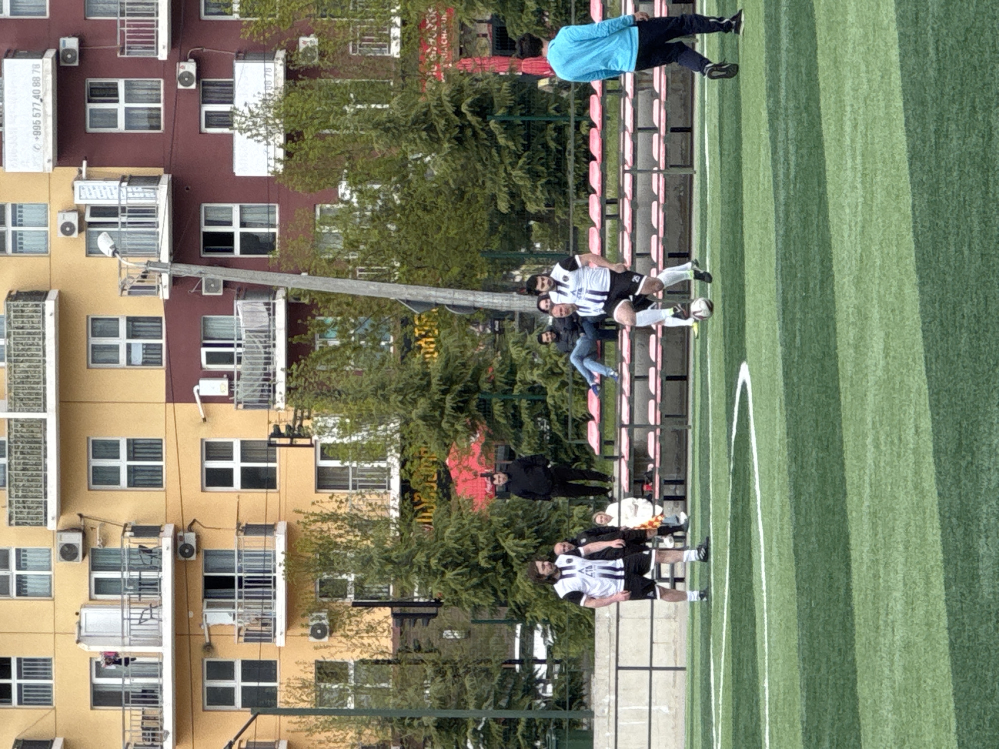 A soccer game is in progress on a green artificial turf field with white lines, adjacent to a set of red stadium seats. Players wearing white and black striped jerseys and black shorts are actively engaged with the ball.
Several individuals are observing the game, including a person in a blue top, a person in a black coat, and other spectators seated in the stadium. The background features an apartment complex with multiple windows and air conditioning units. The buildings are painted in a combination of yellow and red.
The location is likely Tbilisi, Georgia.