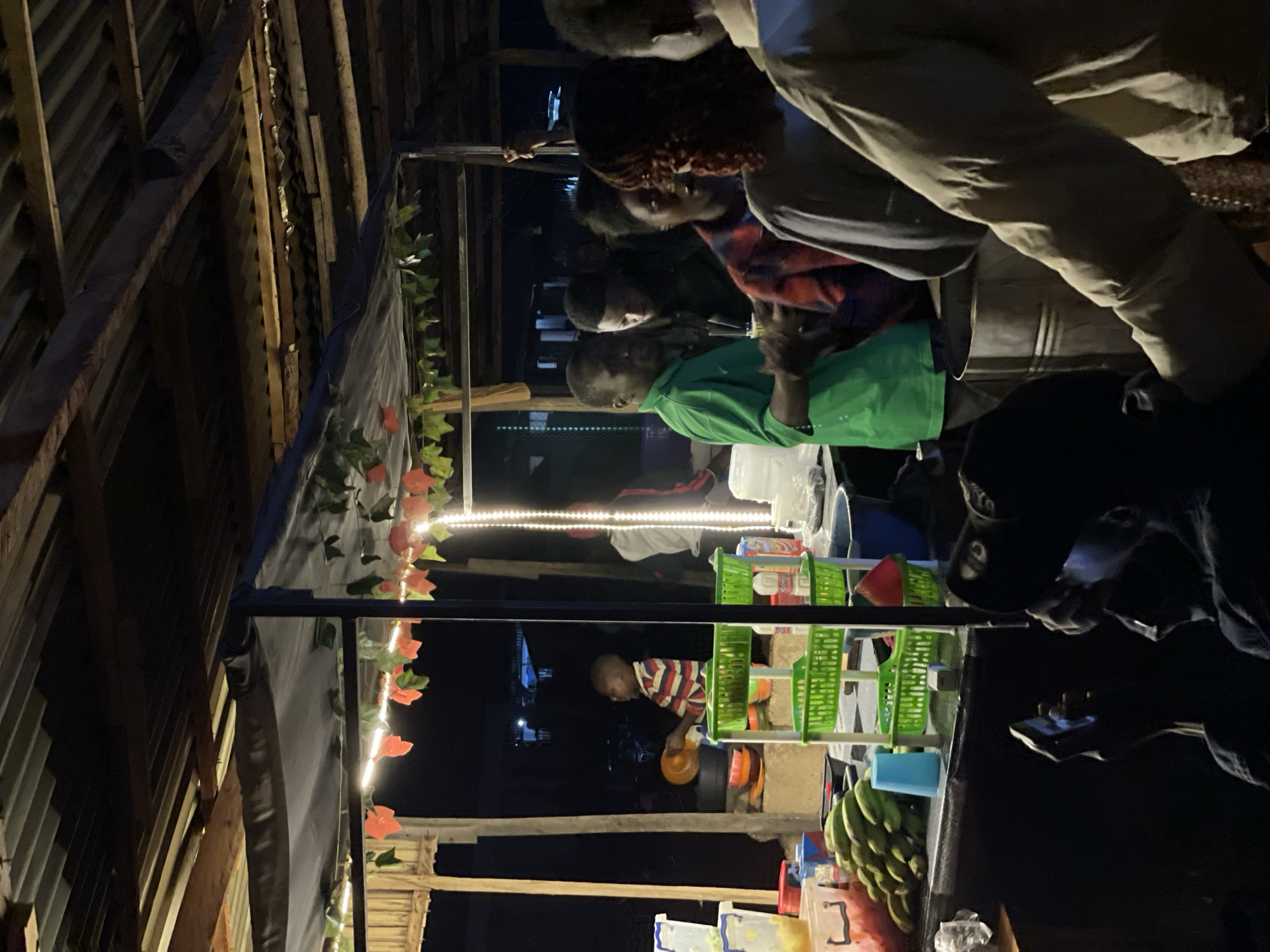 In Durumi, Nigeria, a nighttime street food stall is illuminated by string lights and a bright vertical light. A young boy in a striped shirt is seen holding an orange plate, standing near a stack of colorful bowls and green plastic baskets filled with food items. To his right, a man in a green shirt is interacting with others. In the foreground, a person holds a phone, seemingly capturing the scene. The stall features a variety of produce, including a bunch of bananas. The environment appears to be an outdoor market or roadside setup.