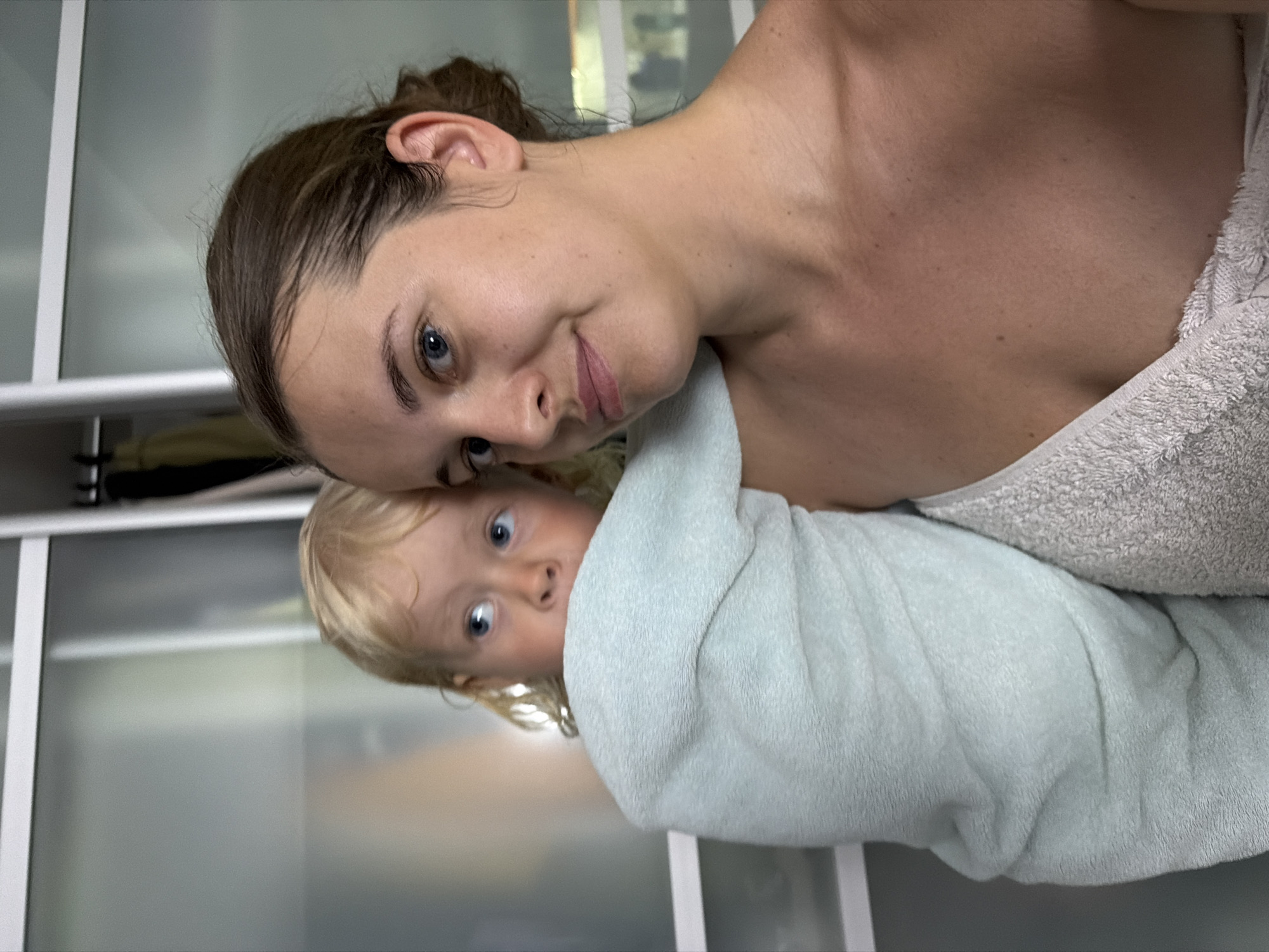In this close-up, selfie-style image, a woman with brown hair pulled back and blue eyes is holding a young child wrapped in a light blue towel. The child, with blonde hair and blue eyes, is looking off to the left. Both are wrapped in towels. The woman is looking at the camera with a slight smile, and the child appears to be peeking from behind her. The background consists of a closet with frosted glass doors and hanging clothes. The image was taken indoors, likely in a residential setting, possibly in Sochi, Russia. The lighting is natural and soft.