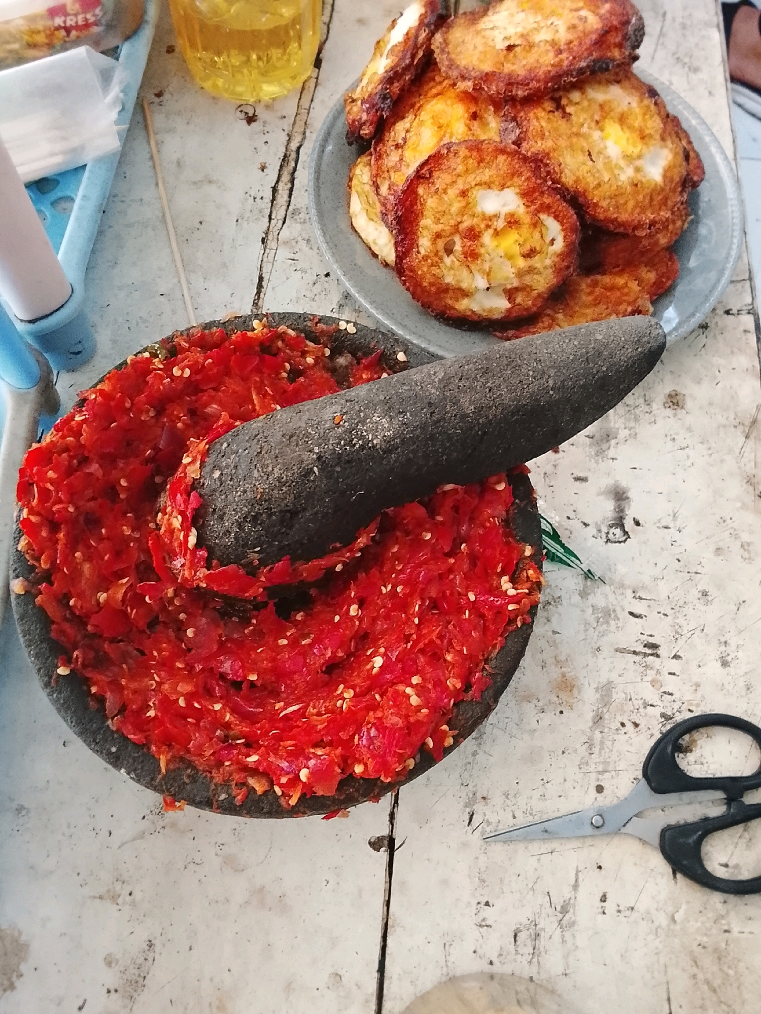 A mortar and pestle filled with a red chili paste sit on a distressed wooden table. To the right of the mortar, a pair of black-handled scissors lies open. In the background, a plate of fried eggs, a glass of yellow liquid, and a blue plastic tray containing a white object and a small packet are visible. The scene suggests food preparation. The photograph appears to be taken in Slawi, Indonesia, given the presence of local ingredients and typical table settings.