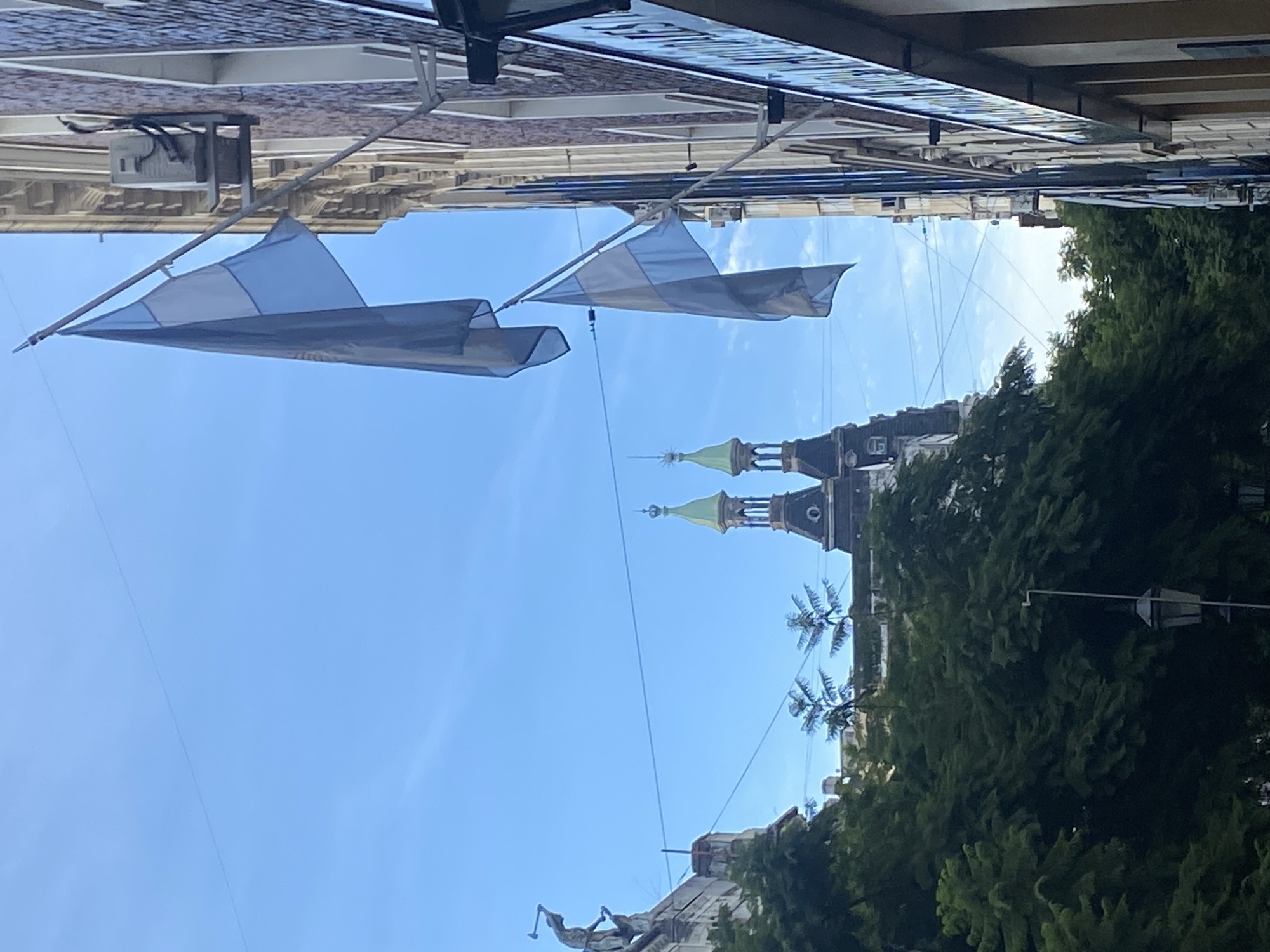 The image shows a street view in Buenos Aires, Argentina. Two light-blue and white Argentine flags hang between buildings. In the distance, the green-topped towers of the Basilica of Our Lady of the Rosary and Saint Dominic are visible above trees. A statue, possibly of a figure on horseback, sits atop a building. Overhead cables crisscross the sky. Buildings line the street with varying architectural styles. The sky is clear and blue. The image orientation is rotated counter-clockwise by 90 degrees.