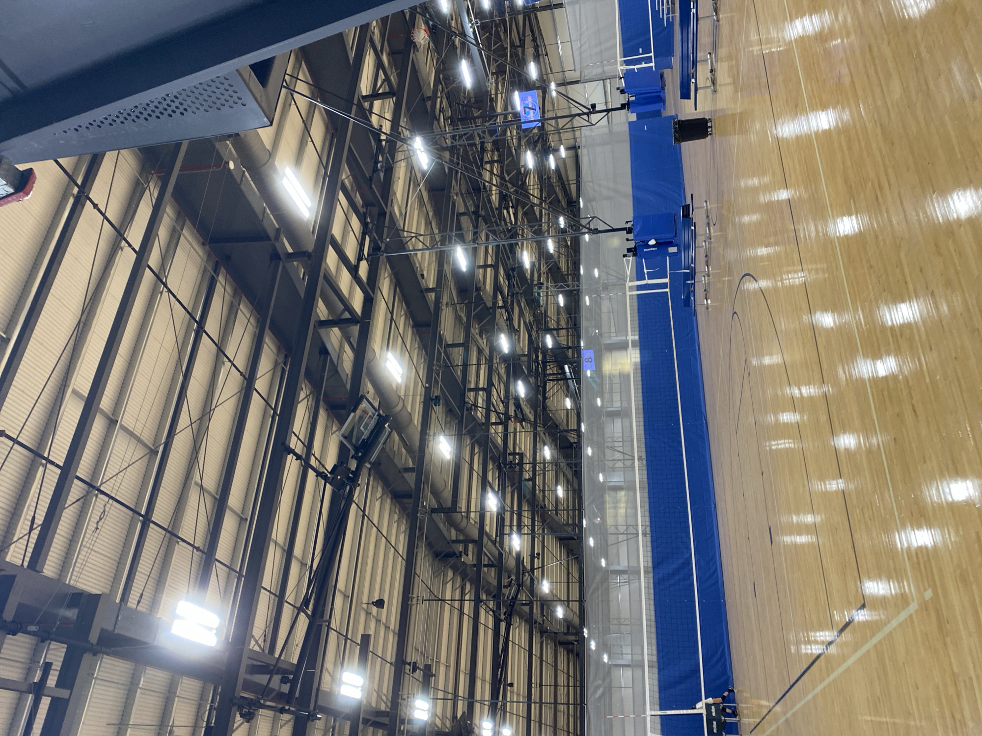 The image shows the interior of a large gymnasium with a wooden floor. The gymnasium is equipped for volleyball, with nets and poles visible. Blue padding is around the poles, and blue curtains hang behind the nets. The ceiling is high and industrial, with exposed beams and ductwork. Numerous fluorescent lights illuminate the space. In the foreground, the polished wooden floor reflects the overhead lights. Along the side of the court, there are bleachers and a trash can. A scoreboard with the number "7" is visible in the distance. The scene appears to be unoccupied by athletes or spectators. The overall impression is of a well-maintained sports facility.