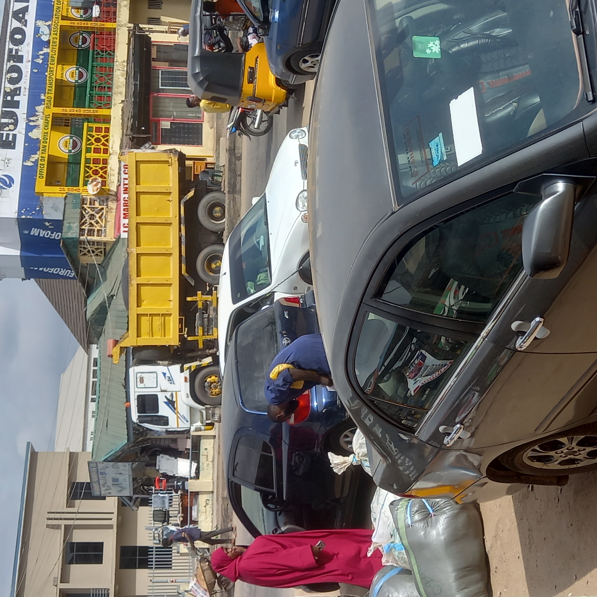 The image shows a street scene in Dadi Riba, Nigeria. Several vehicles are visible, including a yellow dump truck, a white sedan, a blue car being worked on by a person in a blue shirt, a yellow motorized tricycle, and a dark gray sedan. A person wearing a red robe and head covering stands near several large bags wrapped with blue rope. In the background, a building with a sign that reads "EUROFOAM" is visible. Another sign reads "OFFICE OF YAN DOYA CHAPEL ROAD TRANSPORT EMPLOYER'S ASSOCIATION OF."