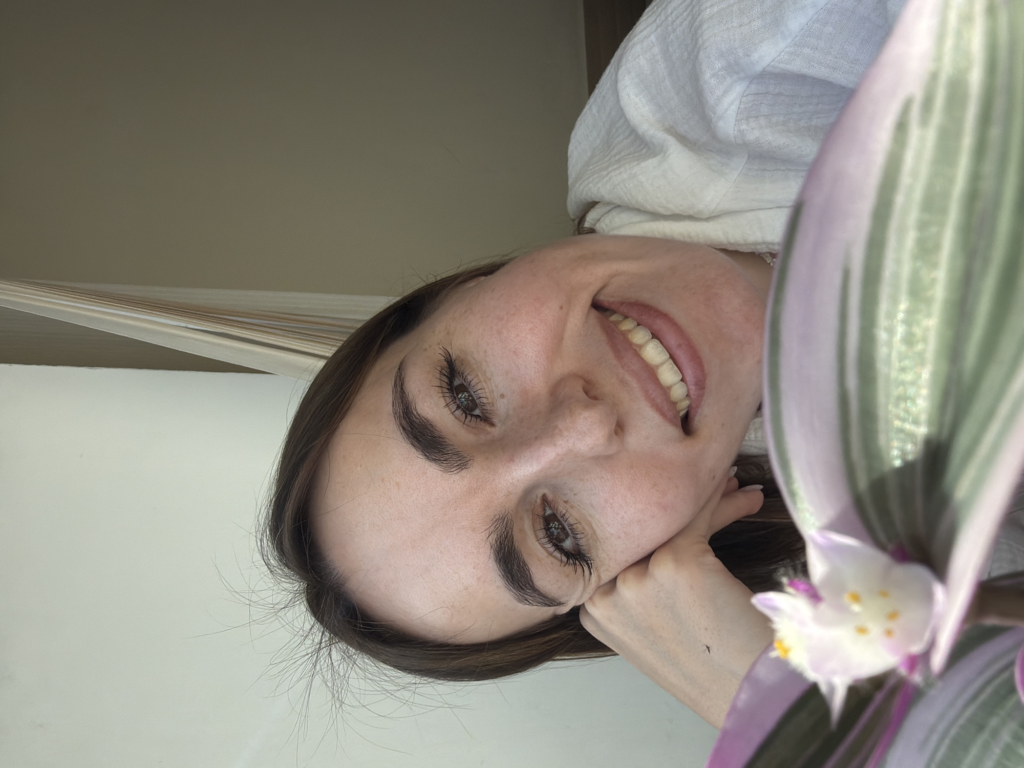 A young woman with light skin, brown hair, and freckles is smiling with her hand resting under her chin. She is wearing a white top. In the foreground, a plant with green and purple striped leaves and a small white flower is visible. The background is a light beige wall and a partial view of a white curtain or screen. The scene appears to be indoors. There is no location in Izhevsk, Russia visible in the image.