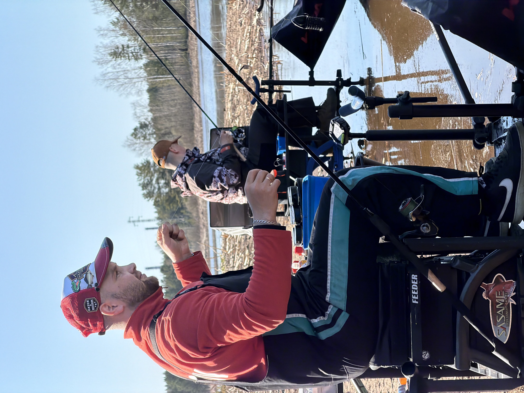 Two men are fishing from platforms on the edge of a body of water, likely a reservoir or lake. The man in the foreground is wearing a red long-sleeved shirt, a cap with patches, and black and teal track pants. He holds the fishing line with both hands. A "Feeder" label and a "Sasame" logo are visible on his fishing platform. In the background, another man sits further back, wearing a camouflage-patterned jacket and a brown cap. The environment includes a body of water with exposed brush along the edge, and bare trees in the background against a clear sky. The location is in or near Chaykovsky, Russia, based on the "Sasame" logo which signifies the Sasame Fishing Hooks, commonly found for Fishing Feeder, or carp fishing, in Russia.