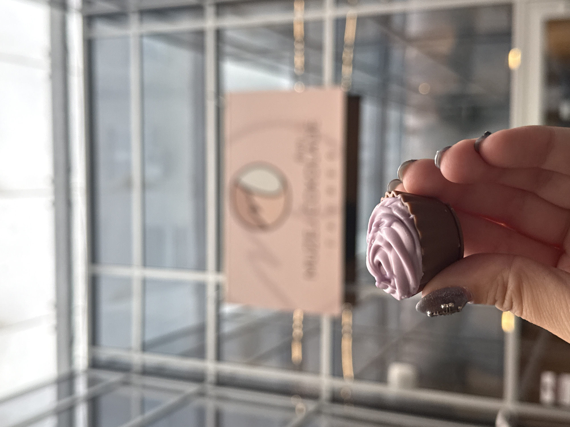 A hand holds a chocolate candy topped with a purple swirl in front of a windowed display case. The case contains a blurred pink sign bearing a logo. The location is identified as being in the city of Zagreb, Croatia, evidenced by the sign. The background includes a structure with large windows. The focus is on the candy and the hand holding it. The person's nails are painted with dark polish and silver glitter accents. The case itself is glass and appears to have a reflective surface.