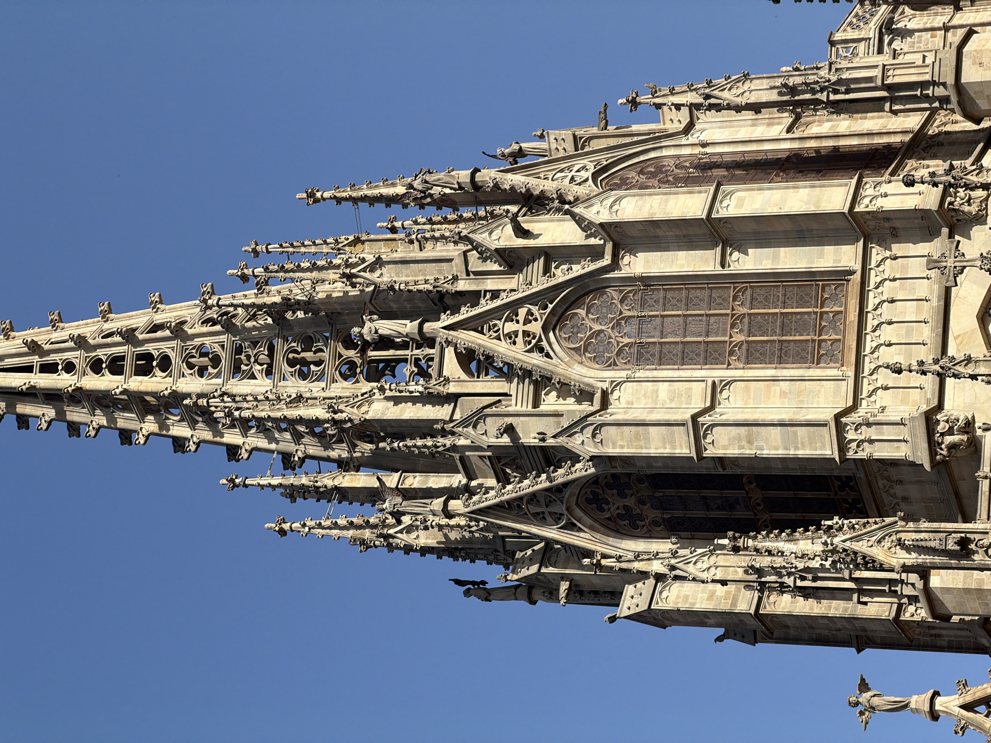 The image shows a close-up of the intricate spires and Gothic architecture of the Barcelona Cathedral. The detailed stonework includes pointed arches, gargoyles, ornate carvings, and statues. Large stained-glass windows are visible on the facade. The sky is a clear, bright blue. The photograph is taken from a low angle, emphasizing the height and grandeur of the cathedral.