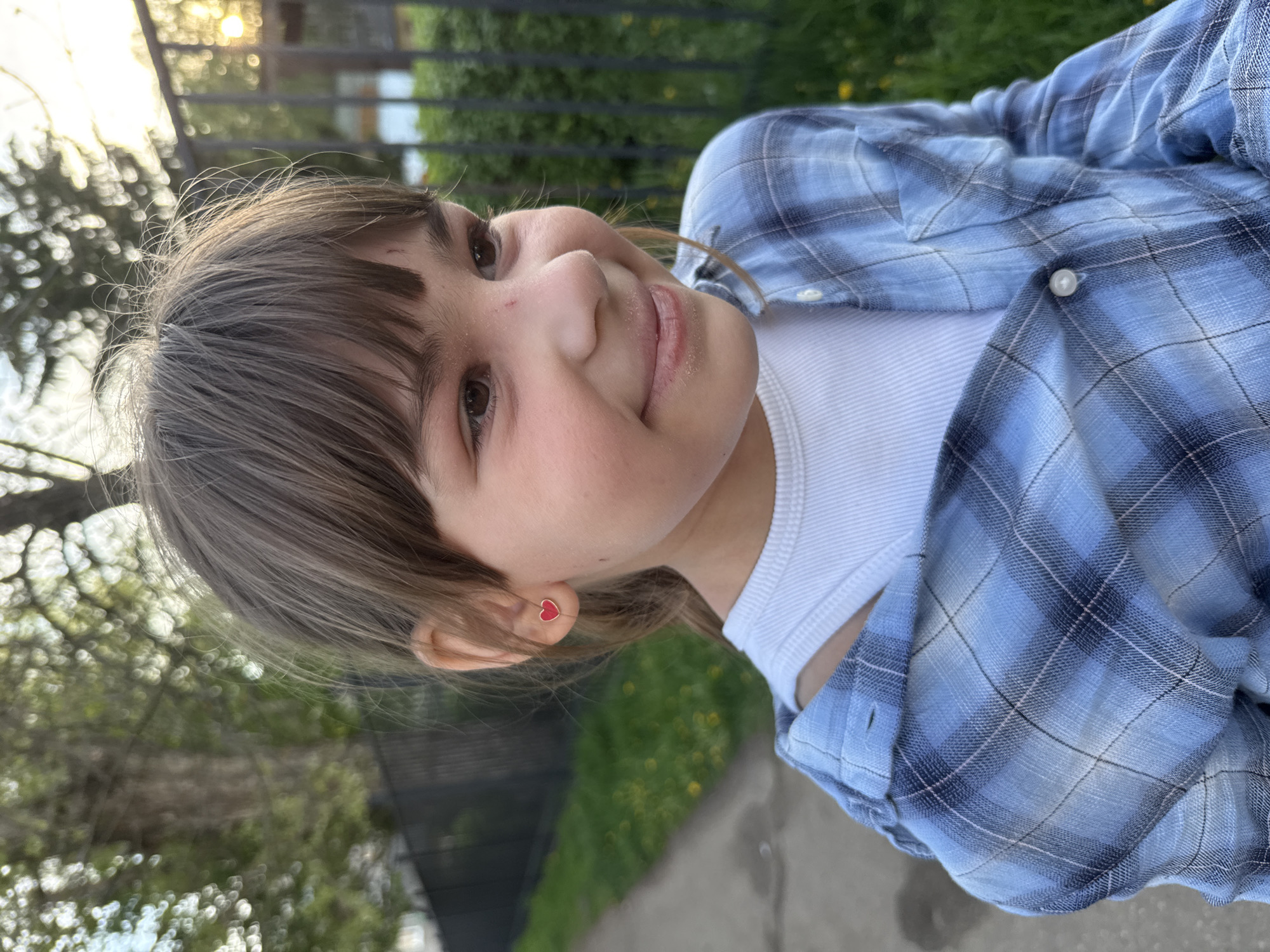 A young girl with brown hair and brown eyes is smiling. She is wearing a white undershirt and a blue plaid shirt. She has a red heart earring in her left ear. The background includes trees, a fence, and a concrete sidewalk. The location is likely in a park or residential area. Given the request, the Rusanovo, Russia, area may be implied, but no details in the image explicitly confirm this.