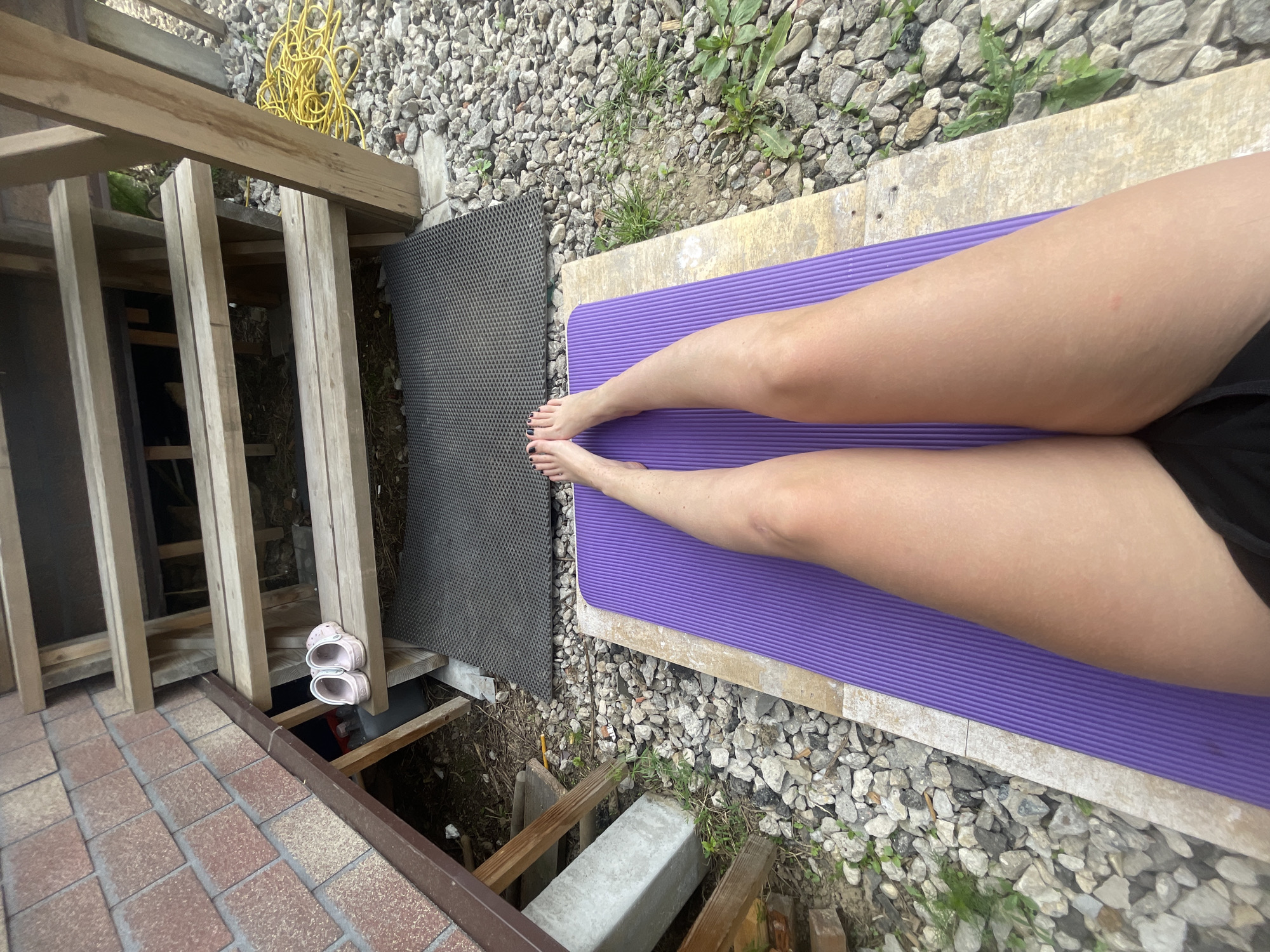 A person lies on a purple yoga mat placed atop a concrete slab surrounded by loose gray gravel. The person's legs are visible, extending from the center of the mat, with the person wearing black shorts. A gray rubber mat is adjacent to the concrete slab and gravel. To the left, there is a wooden structure with visible supporting beams and a set of pink sandals placed on a ledge. Below the wooden structure, there's an unfinished space filled with debris and wooden supports. A section of brick paving is visible in the lower left corner. A yellow electrical cord is scattered among the gravel near the wooden structure. The setting appears to be an outdoor space adjacent to a building or structure. The city is Мосметаллоконструкция Онт, Russia.