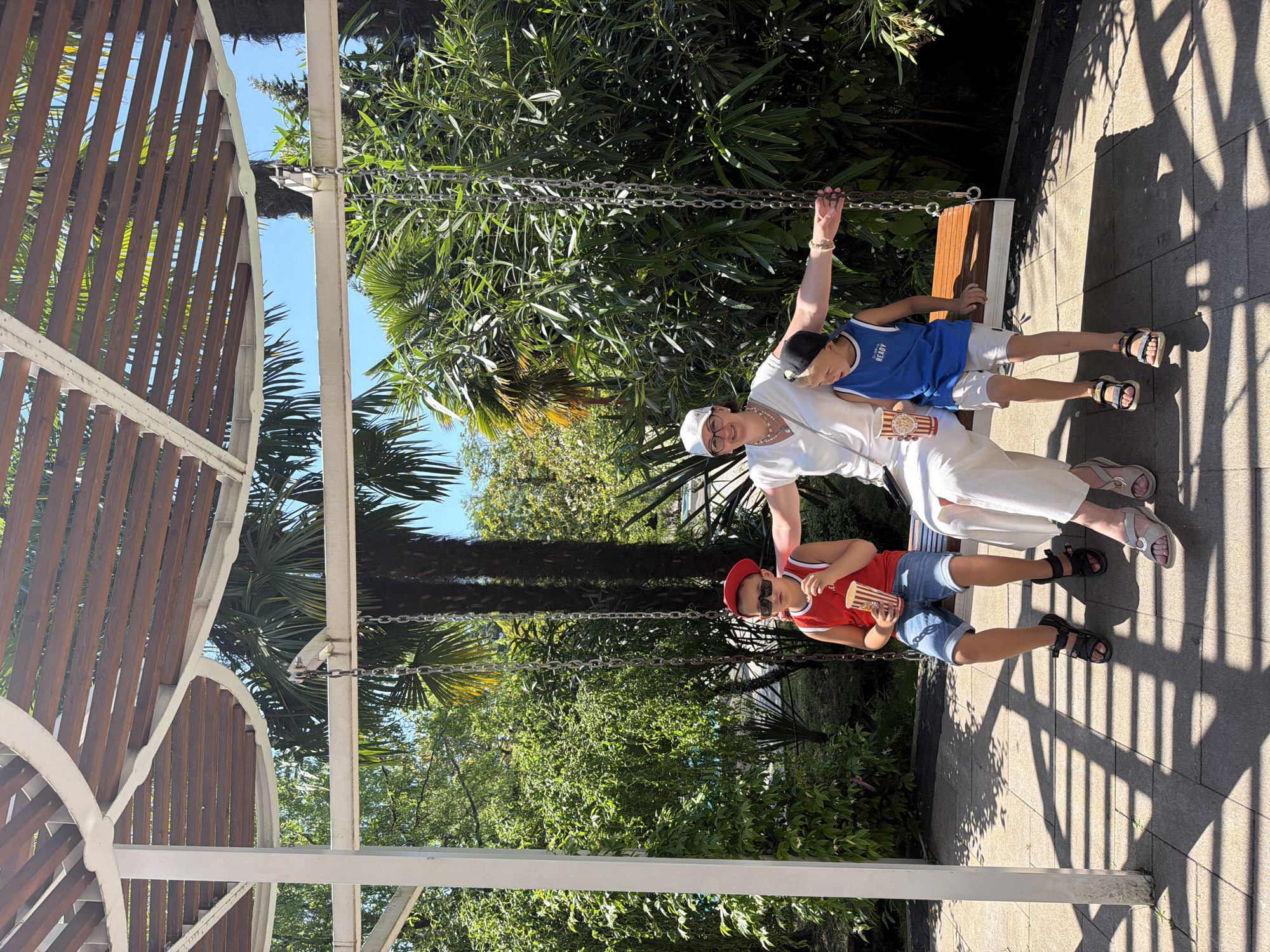 A woman and two young boys are seated on a wooden swing beneath a white and brown pergola. The woman, wearing a white cap, glasses, and a white long-sleeved shirt, is in the center, with a boy on either side. The boy on the left wears a red tank top, denim shorts, a red cap, and sunglasses, holding a cup of popcorn. The boy on the right wears a blue tank top, white shorts, and a black cap, also holding a cup of popcorn. The swing is suspended by chains. The background features lush green foliage, including palm trees, and a clear blue sky. The ground is paved with square tiles, and shadows from the pergola are cast across it. The scene appears to be outdoors in a sunny, tropical or subtropical environment, consistent with the climate of Sochi, Russia.