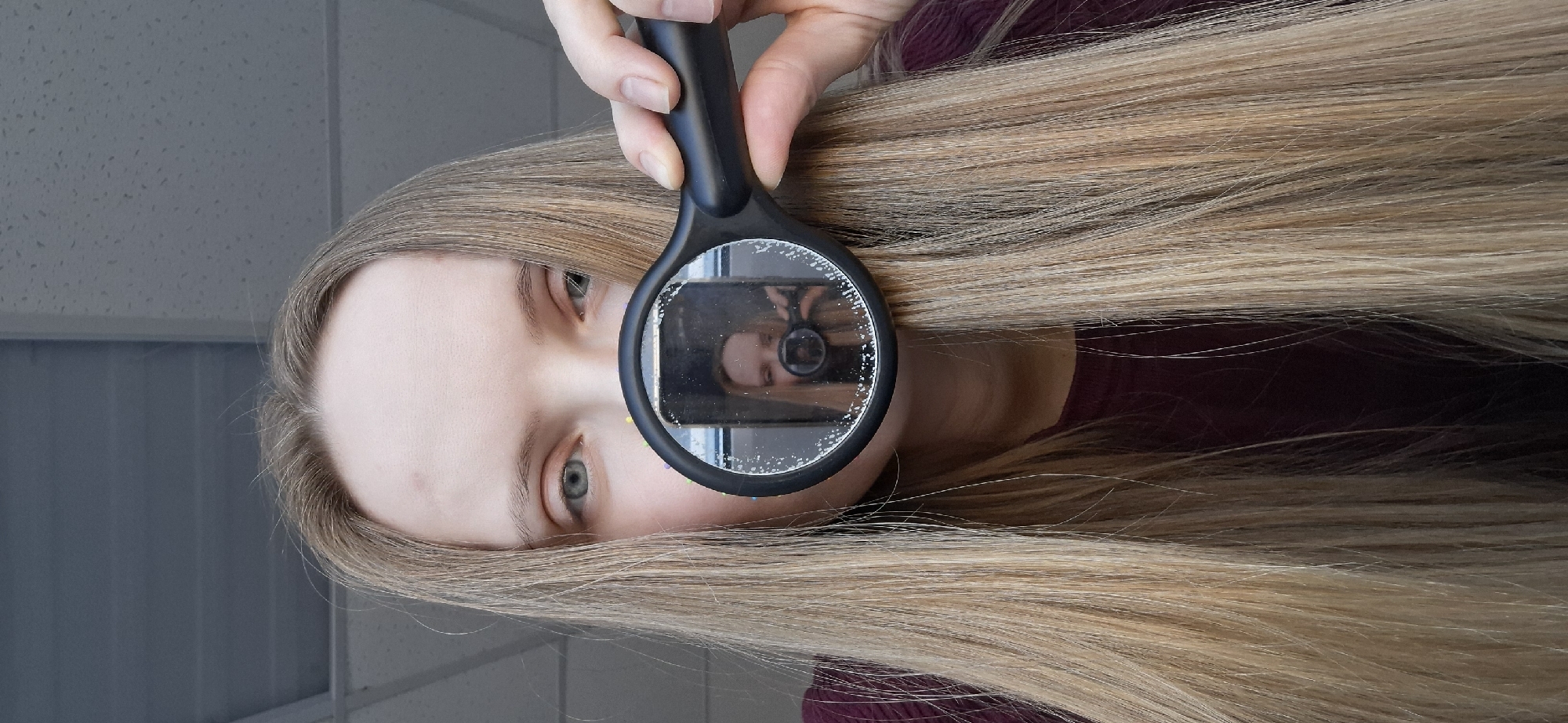 A Caucasian woman is holding a dirty magnifying glass in front of her face. The reflection in the magnifying glass shows her holding a smartphone in her other hand, presumably taking the picture. The woman has blonde hair and light-colored eyes. She is wearing a burgundy top. The background appears to be a room with a light-colored tiled wall. The photo was taken in Chaikovsky, Russia.