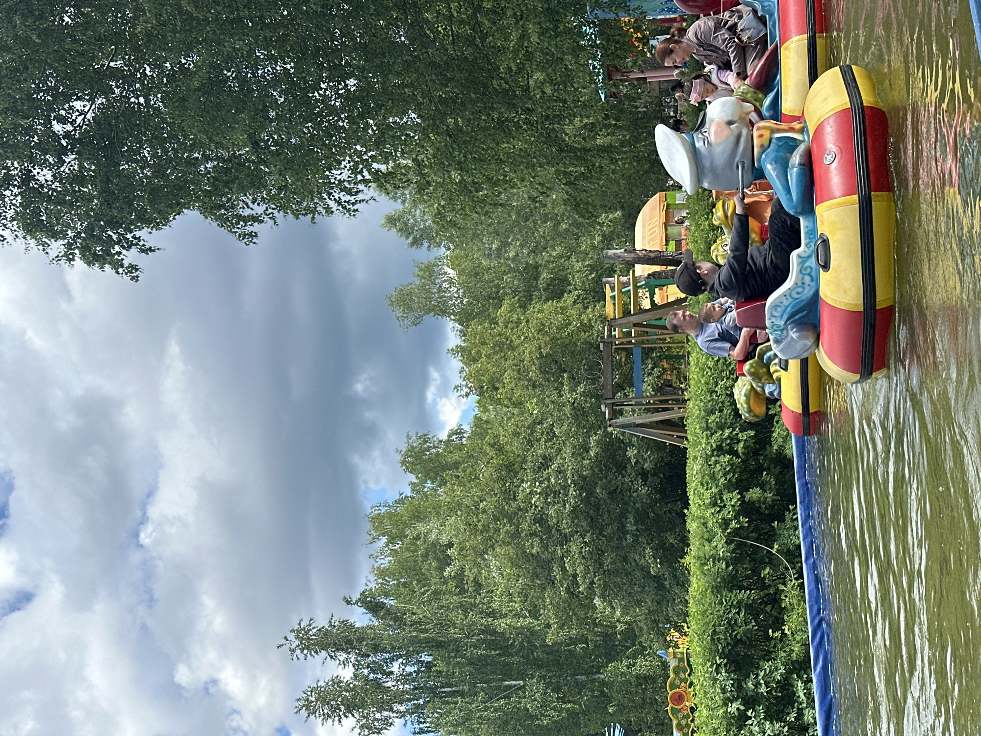 The image depicts a water ride at an amusement park, likely in Chaikovsky, Russia, given the provided context. Multiple boats are visible, each carrying passengers. The boats are brightly colored with red, yellow, and blue elements and appear to be themed with character designs. People of different ages are on the boats, including adults and children. The ride takes place on a body of water, potentially a man-made canal or pool. Lush greenery surrounds the ride, including trees and bushes. The sky is partially cloudy. The overall impression is one of a recreational attraction in an outdoor setting.