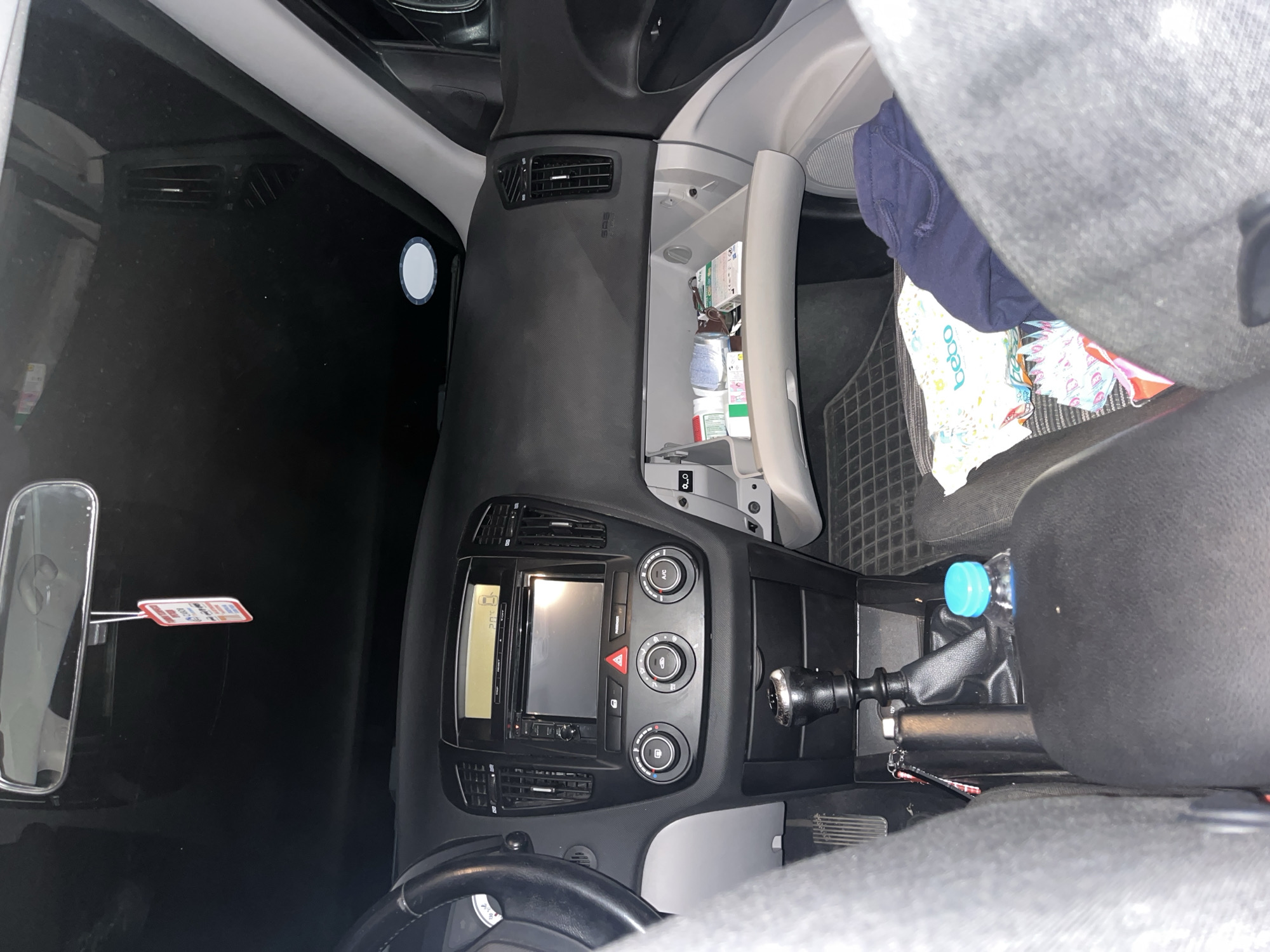 The image shows the interior of a car, specifically the dashboard and front passenger area. The dashboard is black with silver-colored trim and includes a car radio, climate controls, and an open glove compartment filled with various items. A rearview mirror and a parking permit hanging from it are visible. To the right of the dashboard, the front passenger seat is visible, with a water bottle in the cup holder and a baby’s diaper bag on the seat. The location is not discernible from the image, so it is not possible to determine if it is in Marulevo, Bulgaria.