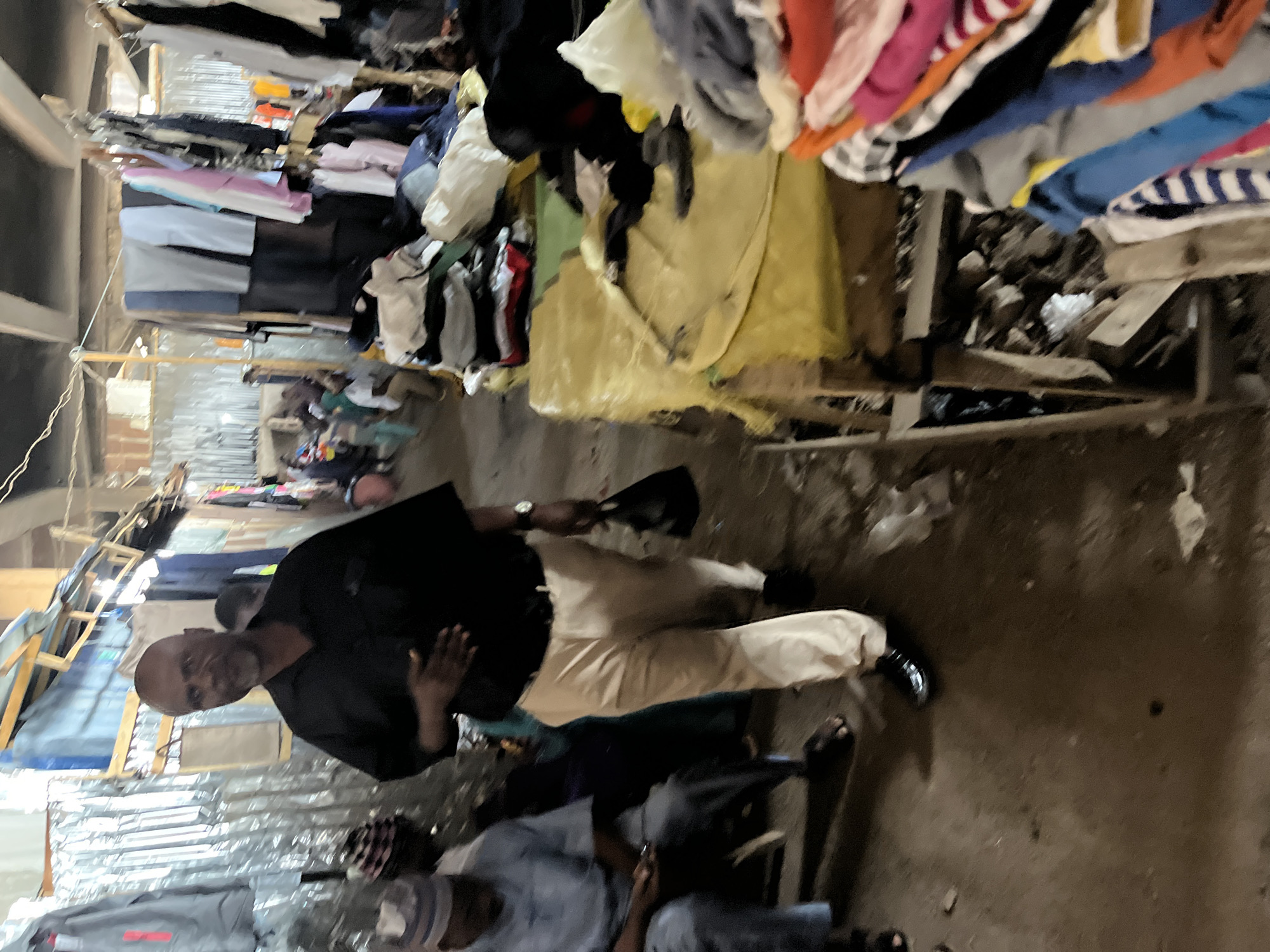 The image depicts a scene in what appears to be a crowded market area in Jos, Nigeria. Overhead, a structure supports racks filled with various items of clothing. A man is seen wearing a black shirt and light tan trousers, holding a black hat, and standing in the aisle of the market. Another man is sitting in a chair, wearing a white top and skull cap. The ground is dusty and cluttered. The environment suggests an informal or semi-permanent commercial setting.