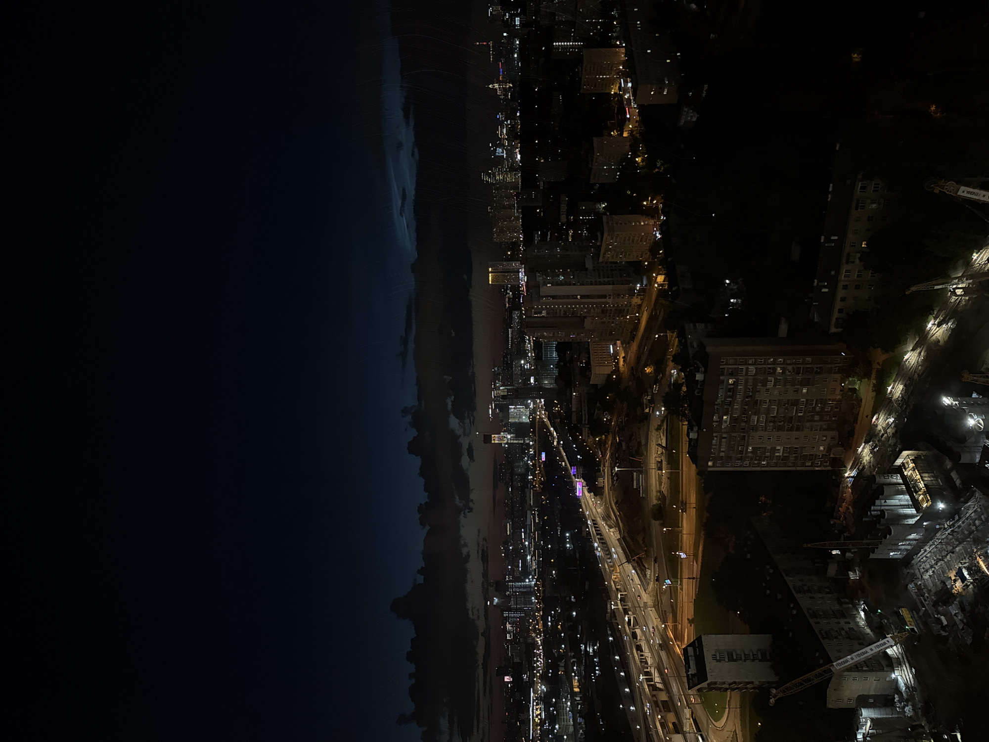 This is an aerial, night-time photograph of a city skyline with a body of water on the left. The image is oriented vertically, with the water on the left and the city on the right. The sky is dark, with some clouds visible over the water, suggesting either dusk or pre-dawn.
The city is illuminated by numerous lights from buildings and street lamps. A highway or major road network is visible in the mid-ground, with cars contributing to the light trails. Several tall buildings and apartment complexes are prominent in the cityscape. On the right side of the image, a construction site is visible with cranes.
The overall mood is one of urban activity and nighttime ambiance. There is no visible text in the image. The image appears to be taken from a high vantage point, likely from another tall building or a drone.