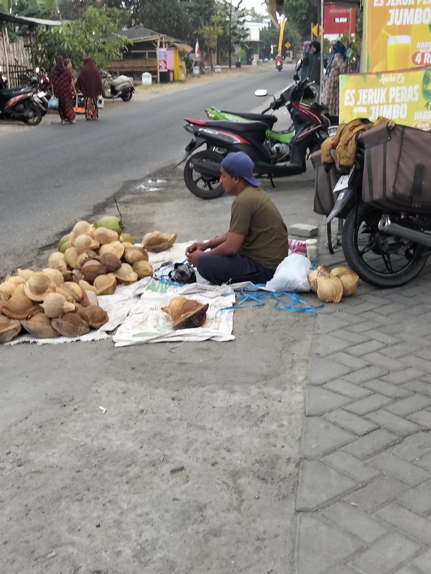 In Gempolan, Indonesia, a man wearing a brown t-shirt and a blue cap sits on the ground next to a pile of halved coconuts and whole coconuts displayed on a white tarp. He is attending to a black bag and some blue rope. Behind him, a motorcycle is parked. In the background, several other motorcycles and people are visible near roadside structures and businesses, with a banner advertising "ES JERUK PERAS JUMBO." The scene appears to be a street-side market or vendor stall.