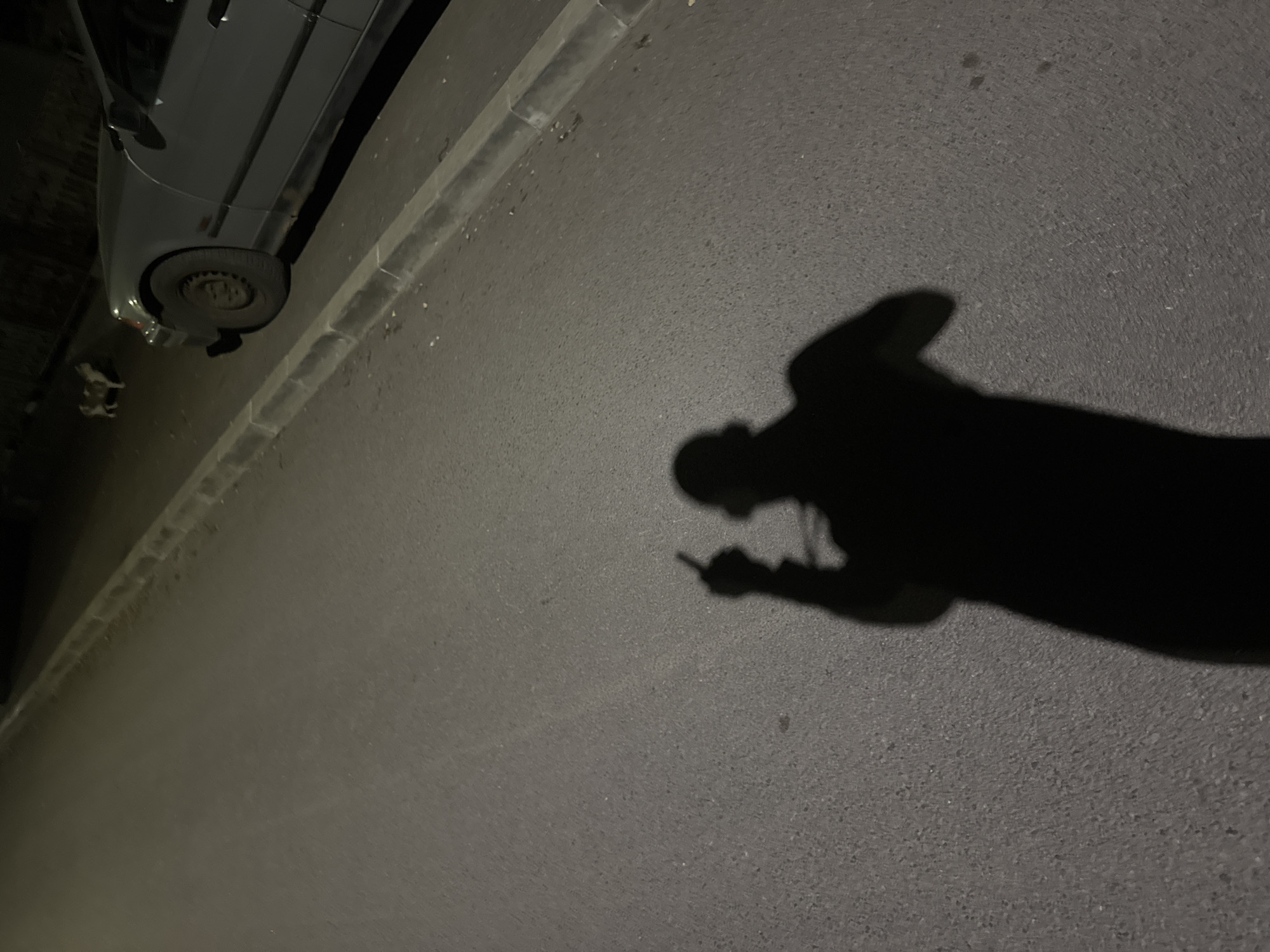 The image depicts an urban street scene at night, likely in Sofia, Bulgaria. The surface is asphalt. A portion of a dark-colored car is visible in the upper left corner. The curb is concrete. The dominant feature is the shadow of a person holding an object that could be a phone or cigarette. The shadow suggests the person is standing on the pavement. Overall the street is not well illuminated.