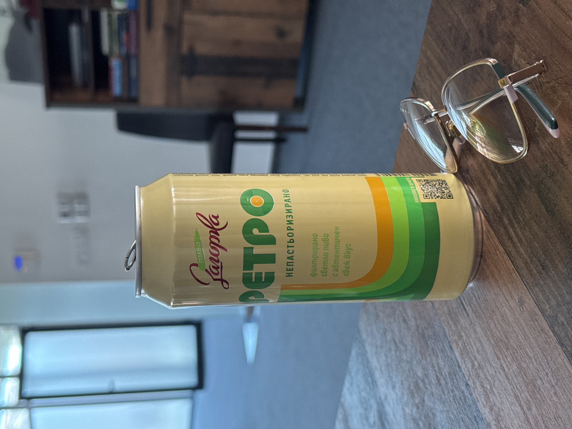 A can of "Пиво Саторка Ретро" (Satorca Retro Beer), a Bulgarian beer, lies on a wooden surface. The can's design features retro graphics in orange, yellow, and green bands. Next to the can, there is a pair of eyeglasses. The background includes a wooden shelving unit and a blurred window or opening, suggesting an interior setting. Based on the beer brand's origin, it can be inferred that the image was taken somewhere in Bulgaria, likely in or near Sofia.