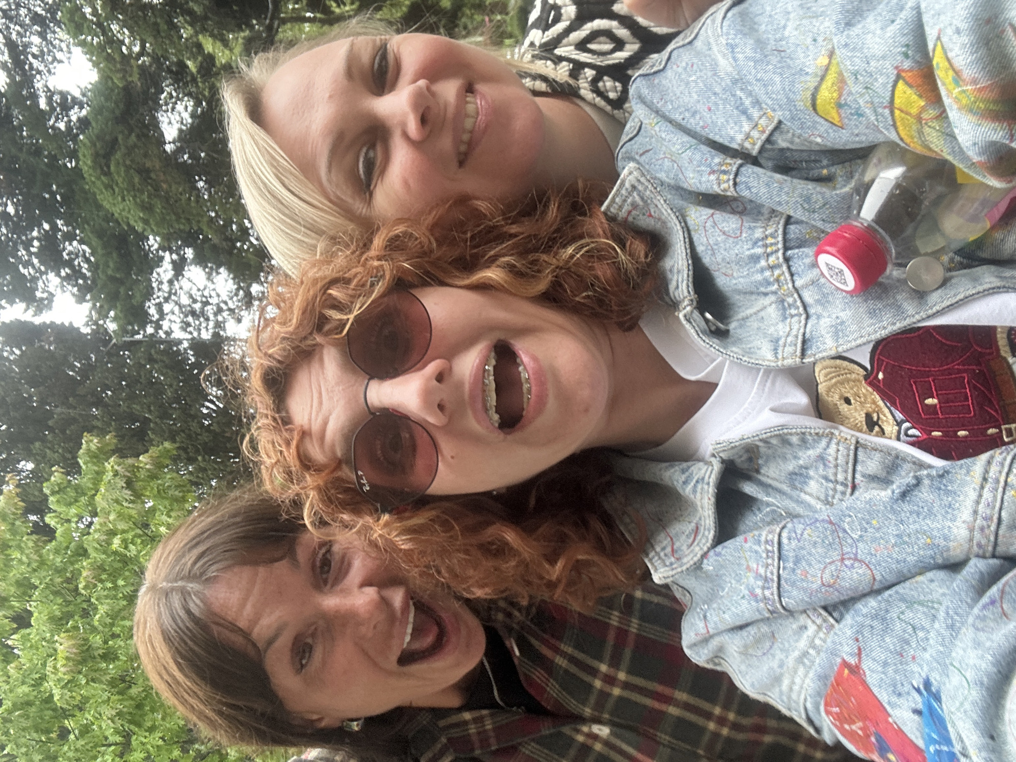 A rotated, close-up, eye-level shot shows three women posing for a selfie outdoors, with trees visible in the background. The woman at the bottom left has short brown hair with bangs and is wearing a plaid shirt. She is smiling and her mouth is open. The woman in the middle has red curly hair and round sunglasses. Her mouth is also open and she is wearing braces. A white T-shirt is visible under a blue denim jacket with colorful paint details. A small, empty bottle with a red cap is tucked into a pocket. The woman at the top right has blond hair and is wearing a black and white patterned shirt. She is smiling. Sochi, Russia, cannot be confirmed based on the visual information provided.