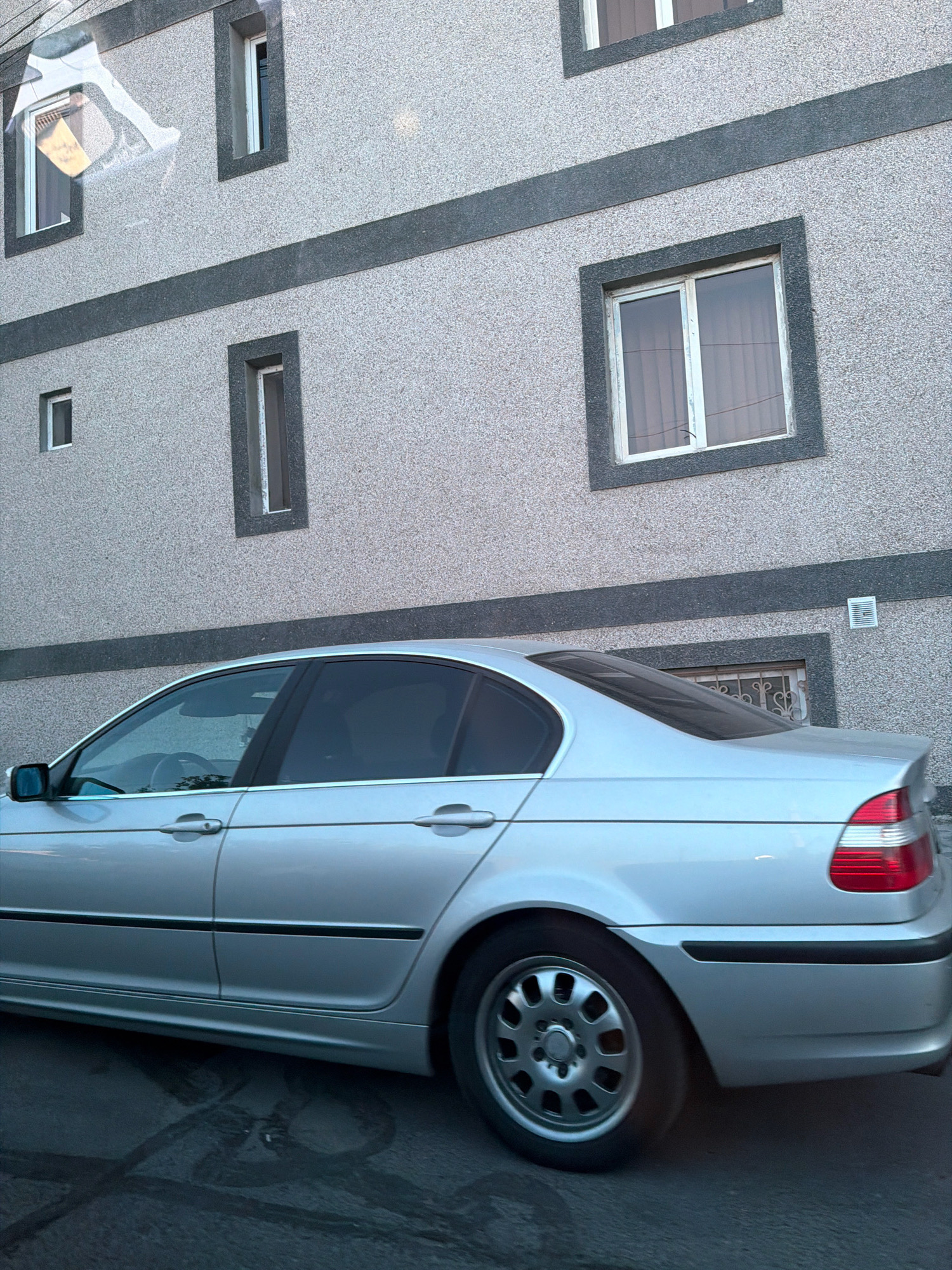 A silver sedan is parked in front of a building with a speckled gray facade in Yerevan, Armenia. The building has horizontal dark gray bands and several windows. The car's side profile is visible, showing its doors, wheels, and rear taillight. The ground is asphalt, with some dark, wet-looking marks. The lighting suggests it is daytime, possibly late afternoon or early morning, given the shadows and reflections.