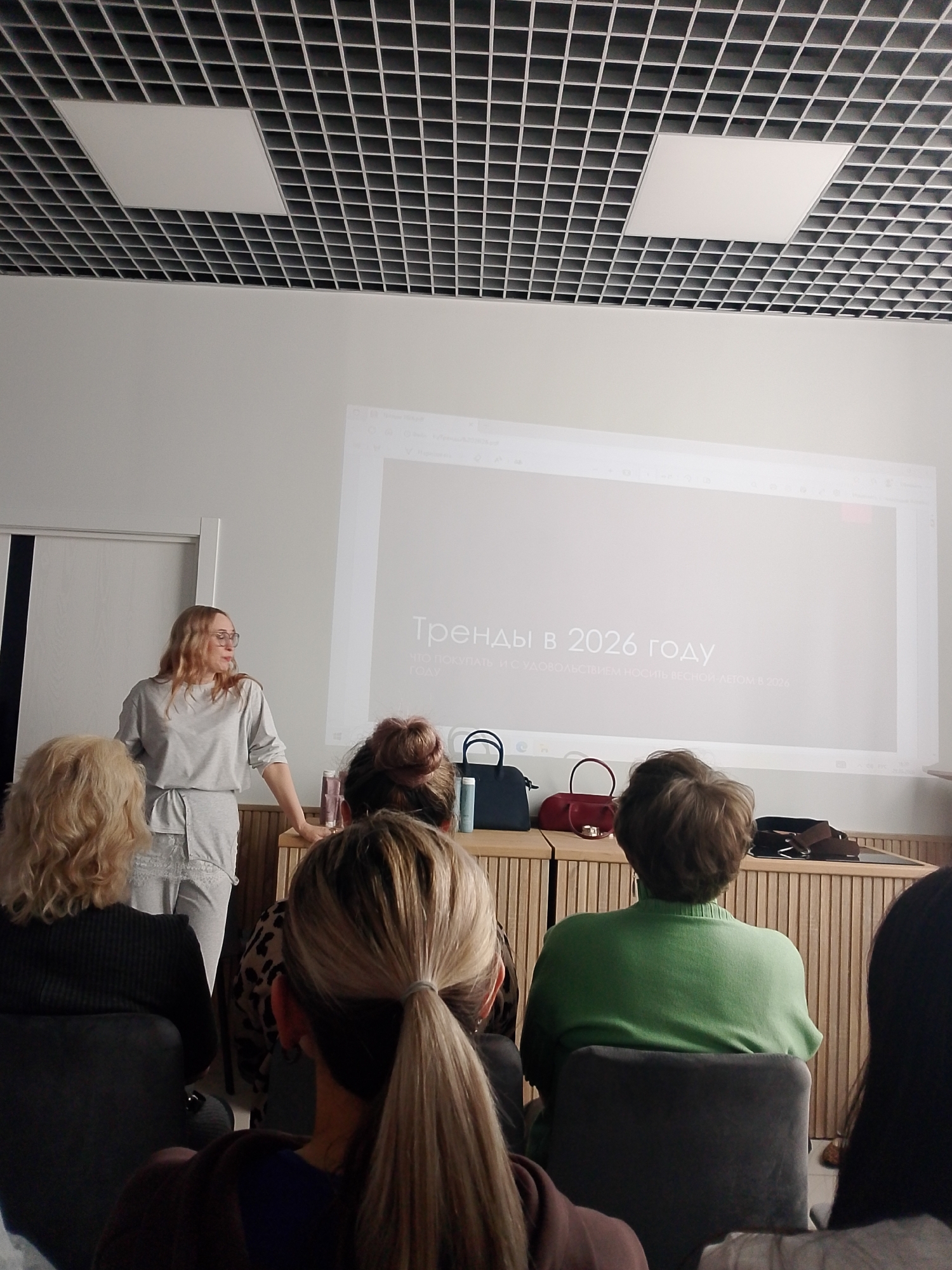 Here's a description of the image:
**Scene:** An indoor setting resembling a conference room or lecture hall. A woman stands at the front, giving a presentation. Several people are seated, facing her, presumably the audience.
**People:**
* A woman with blonde hair and glasses stands presenting. She wears a gray outfit.
* Several audience members are visible, with varying hairstyles and clothing. Their faces are not clearly visible.
**Subjects/Objects:**
* A projector displays a presentation slide in Russian. The heading translates to "Trends in 2026."
* A wooden dresser acts as a backdrop to the presenter, with handbags/purses placed on it.
* There are chairs for the audience to sit on.
* The ceiling has a grid pattern with inset lights.
**Actions:**
* The woman is giving a presentation, gesturing to the screen.
* The audience members are presumably watching and listening to the presentation.
**Environment:**
* The room appears to be well-lit.
* The overall atmosphere is professional or educational.
**Location Details:**
* The text on the projection mentions the Russian language. It is possible that the location is Izhevsk, Russia, but there is no concrete evidence for the city.
