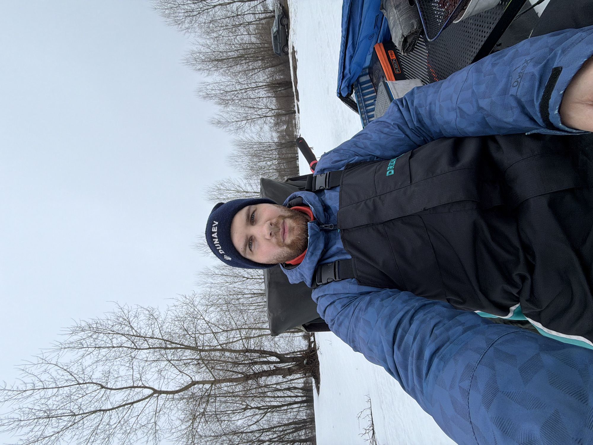A man is outside in a snowy environment near trees. He is wearing a blue hat with white lettering and a black vest over a blue jacket. There is a vehicle and other objects nearby. The location is in Noviy, Russia.