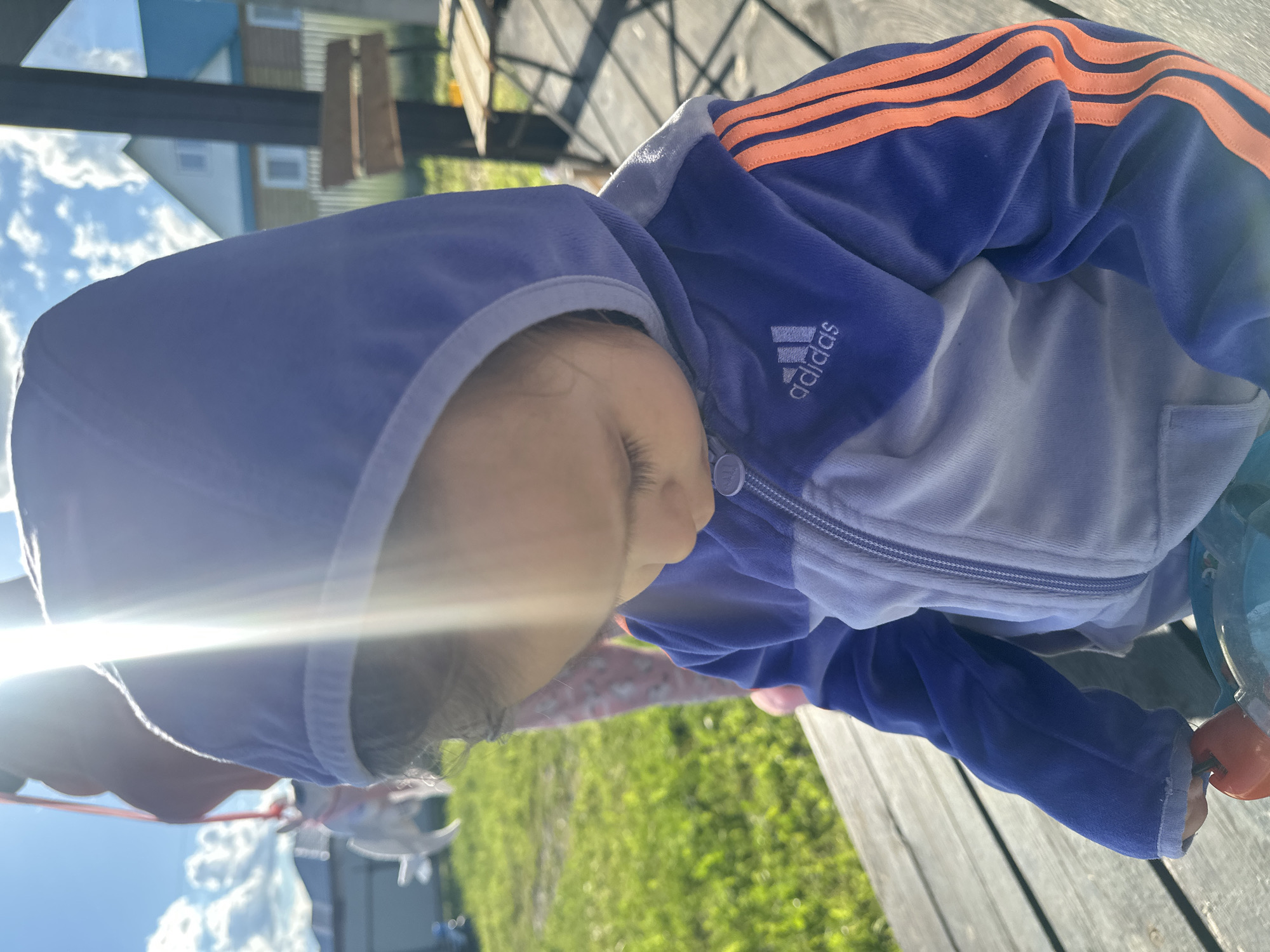 A young child wearing a blue and orange Adidas hooded jacket is partially visible, likely outdoors. The child is facing downward, with their face partially obscured by the hood and strong sunlight. The background shows elements of an outdoor setting, including a wooden deck or platform, grass, and glimpses of buildings and the sky. The exact location in Kharnavy, Russia, cannot be determined from the image alone.