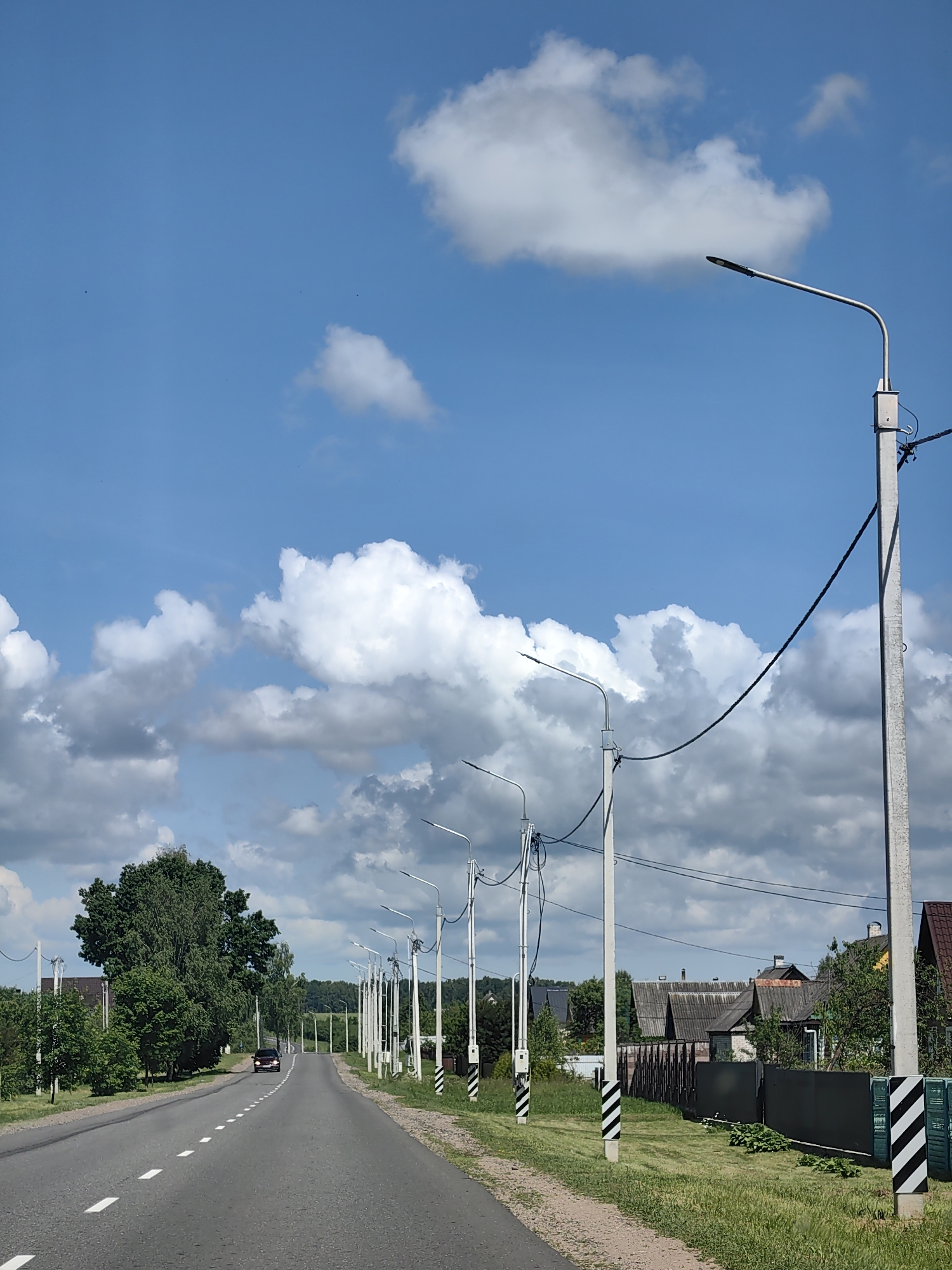 The media file depicts a rural road scene on a bright, partially cloudy day. The road stretches into the distance, lined with numerous streetlights and power poles, suggesting a somewhat developed rural area. On the right side of the road, houses with dark roofs and fences are visible. Lush green trees and grass flank the road, indicating a warm season. The sky is a vibrant blue, dotted with fluffy white clouds. A single car is driving away from the viewer down the road. The scene appears peaceful and tranquil, with no people visible. The time of day is likely daytime, given the bright sunlight and clear sky. The location is identified as Ilyanskiy selskiy Sovet, Belarus.
