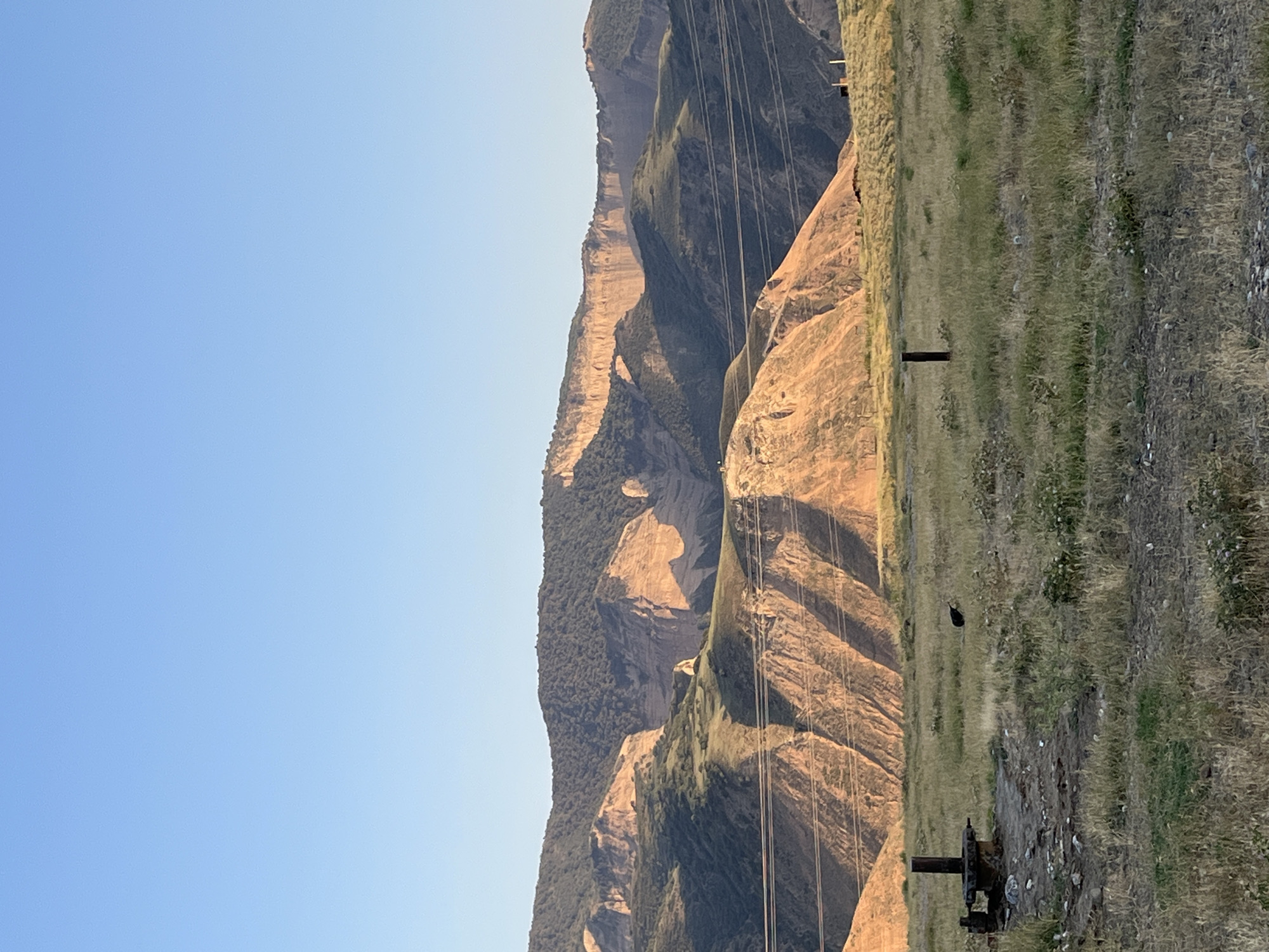 The image depicts a vast, arid landscape dominated by rolling hills and rugged mountains under a clear blue sky. The foreground is characterized by dry, overgrown grass and sparse vegetation. Several objects are visible in the foreground, including what appears to be a rusty, abandoned piece of machinery or wellhead with a vertical pipe, a wooden fence post, and in the middle ground, a dark, bird-like figure. A power line stretches across the mid-section of the image, running horizontally in front of the mountainous backdrop. The mountains themselves exhibit layered rock formations, with slopes covered in a mix of vegetation and exposed earth, catching the warm light of what appears to be late afternoon or early morning sun, casting long shadows. The overall impression is one of a remote, natural environment. The location is identified as Mailuu-Suu City, Kyrgyzstan.