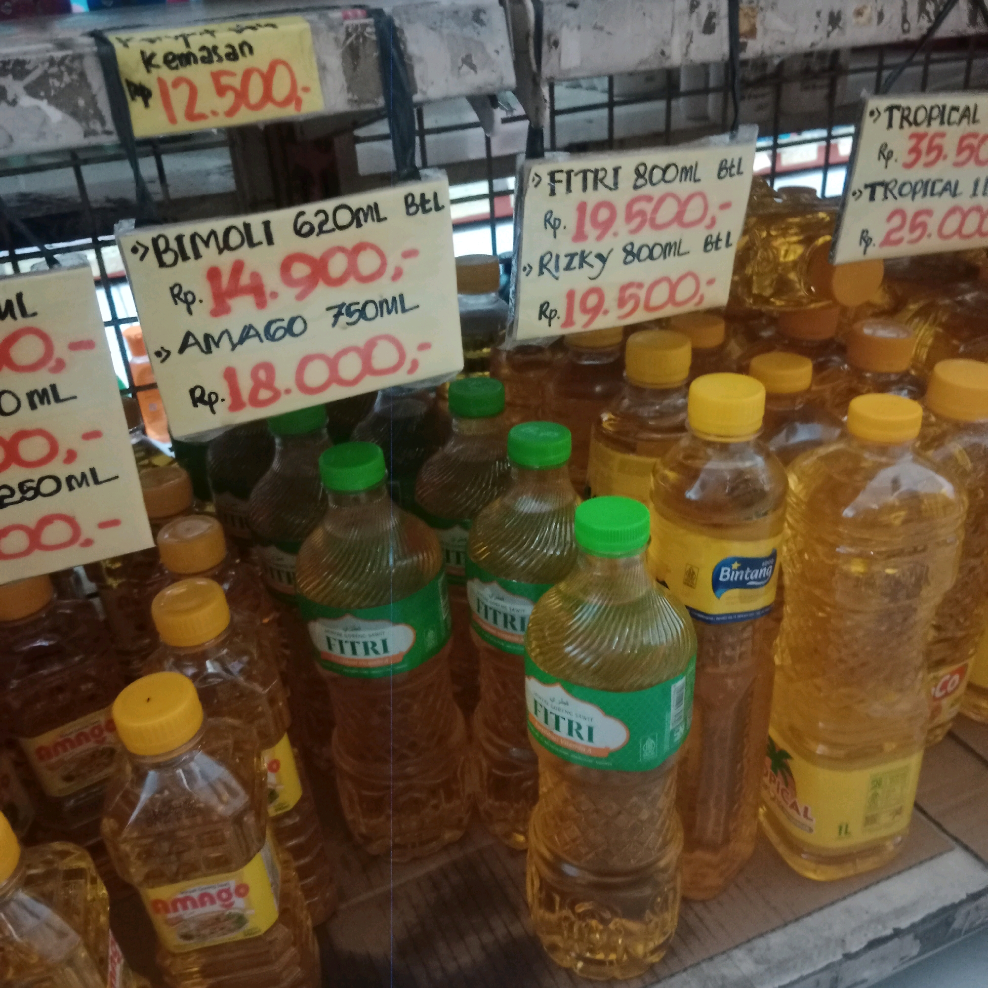 The image shows a display of cooking oil bottles for sale in an outdoor market setting, possibly in Tegowanu Wetan, Indonesia. Several yellow and white price tags are attached to the bottles. One price tag indicates "kemasan Rp. 12.500,-". Another tag reads ">>BIMOLI 620ML BTL Rp. 14.900,-". Below that, a tag says "AMAGO 750ML Rp. 18.000,-". Further to the right, a tag reads ">>FITRI 800ML BTL Rp. 19.500,-", and below it, ">>RIZKY 800ML BTL Rp. 19.500,-". On the far right, a price tag is partially visible with ">>TROPICAL" and "Rp. 35.50" and another below it reads ">>TROPICAL 1L Rp. 25.000,-". The bottles are filled with a yellow liquid, presumably cooking oil. Some bottles have green caps, while others have yellow caps. One bottle prominently displays the brand "Bintang". The bottles are arranged on a wooden surface. In the background, a metal grate structure is visible. The lighting suggests it is daytime, and the overall scene appears to be a local market stall. There are no people clearly visible in the frame.