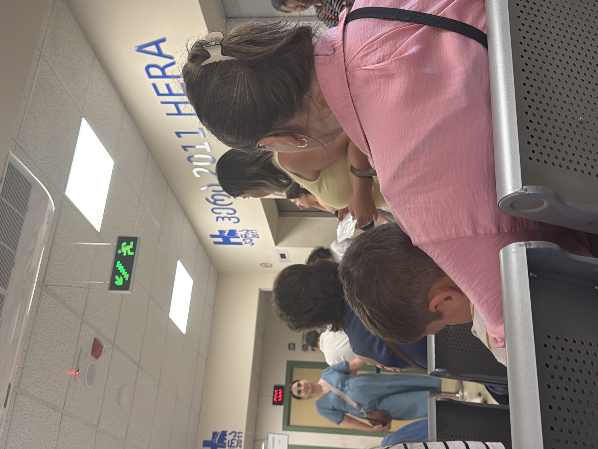 The image depicts the interior of a waiting area, possibly a medical facility, in Tbilisi, Georgia. The walls are painted a light beige, with blue lettering that reads "2011 HERA" and a blue stylized "H" symbol, suggesting it is a branch of HERA (National Center for Disease Control and Public Health) or a similar organization established around 2011. Fluorescent ceiling lights illuminate the space, and a green illuminated exit sign with figures indicating stairs and walking is visible on the ceiling. In the foreground, a young child, viewed from behind, sits in a perforated gray chair, with their head resting on the shoulder of a person wearing a pink shirt. To the left, a woman in a blue dress with a crossbody bag is walking. Other individuals are visible in the background, suggesting a waiting room environment. The atmosphere appears calm and orderly.