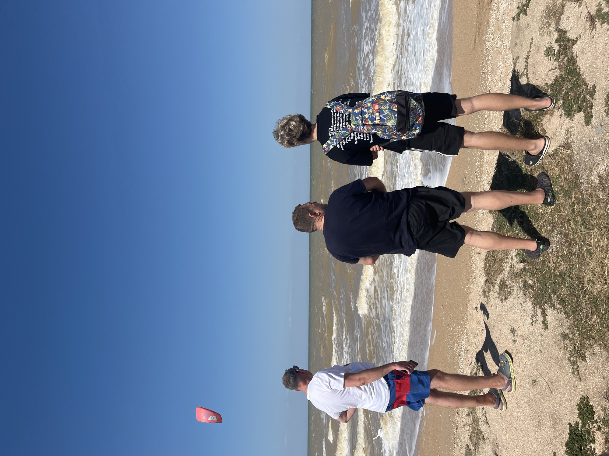 Three individuals are observed from behind, standing on a sandy beach facing a body of water under a clear blue sky. A red kite is visible in the upper left quadrant of the image. The individual on the left wears a white t-shirt and red and blue patterned shorts, with grey and yellow sandals. The middle individual wears a black t-shirt and black shorts, with black slip-on sandals. The individual on the right wears a black t-shirt with a colorful, patterned backpack and black shorts, with black and white slip-on sandals. The sea is moderately choppy with waves breaking on the shore. The location is the beach in Dolzhanskaya, Russia.