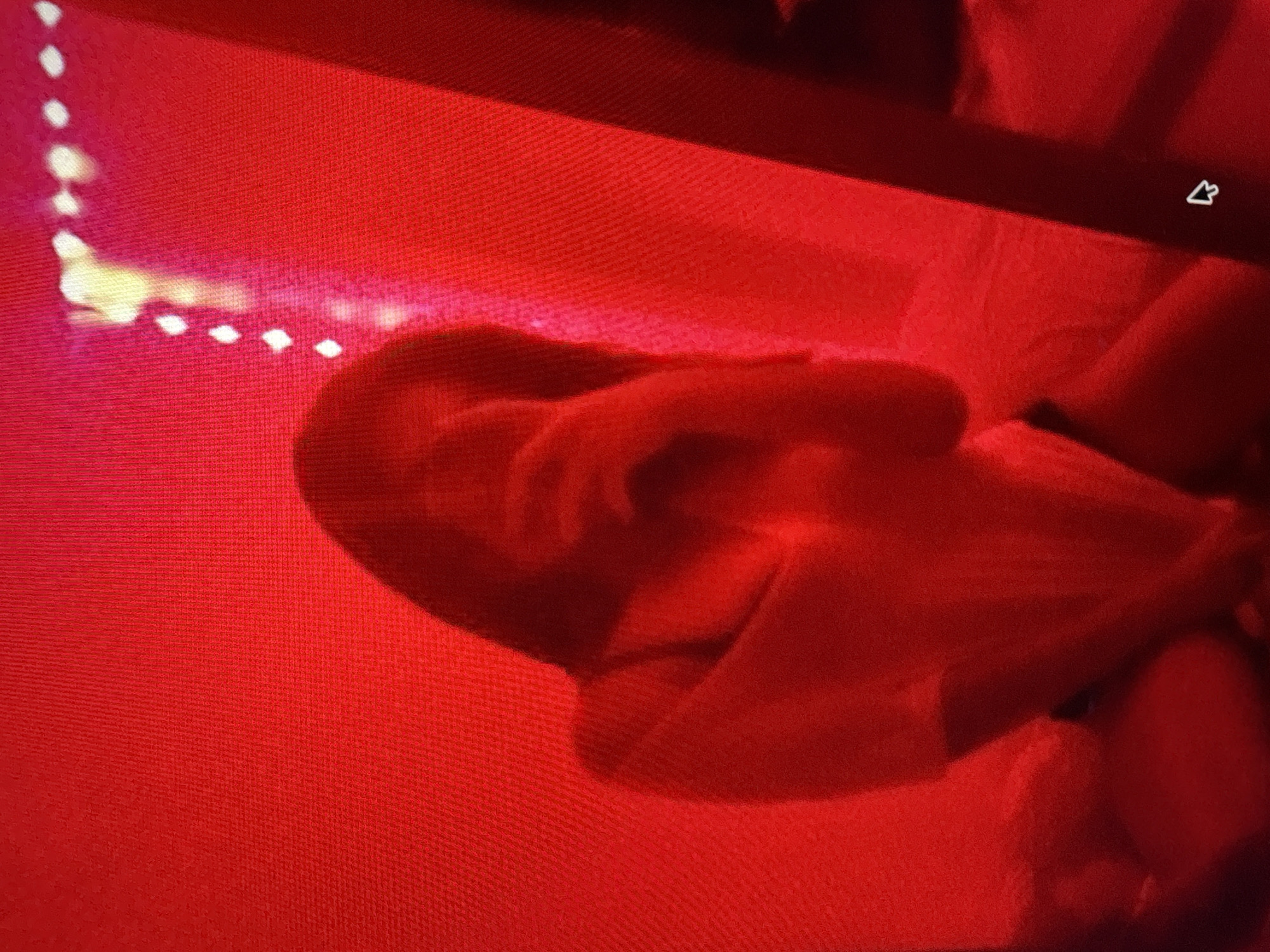 The image shows a person's torso and head, illuminated by red light. The person is wearing a light-colored shirt. The background is a solid red color, with some faint vertical lines visible. There are also some bright, blurry lights in the upper right portion of the image, which could be part of a mirror or a decorative light fixture. The overall impression is of a dimly lit room with a strong red hue.