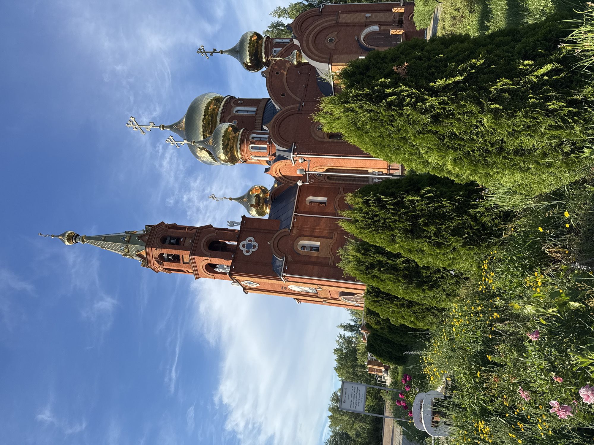 The image shows a large, red brick Orthodox church with multiple golden domes and a tall bell tower. The church is surrounded by a lush green landscape featuring manicured cypress trees, a variety of flowers in bloom (including pink peonies and yellow wildflowers), and a patch of grass. In the foreground, there are two white benches. A signpost with Russian text is visible to the left of the church. The sky is blue with scattered white clouds. The scene appears to be tranquil and well-maintained. This is the Holy Trinity Church (Svyato-Troitskiy Khram) in Chaikovsky, Russia.