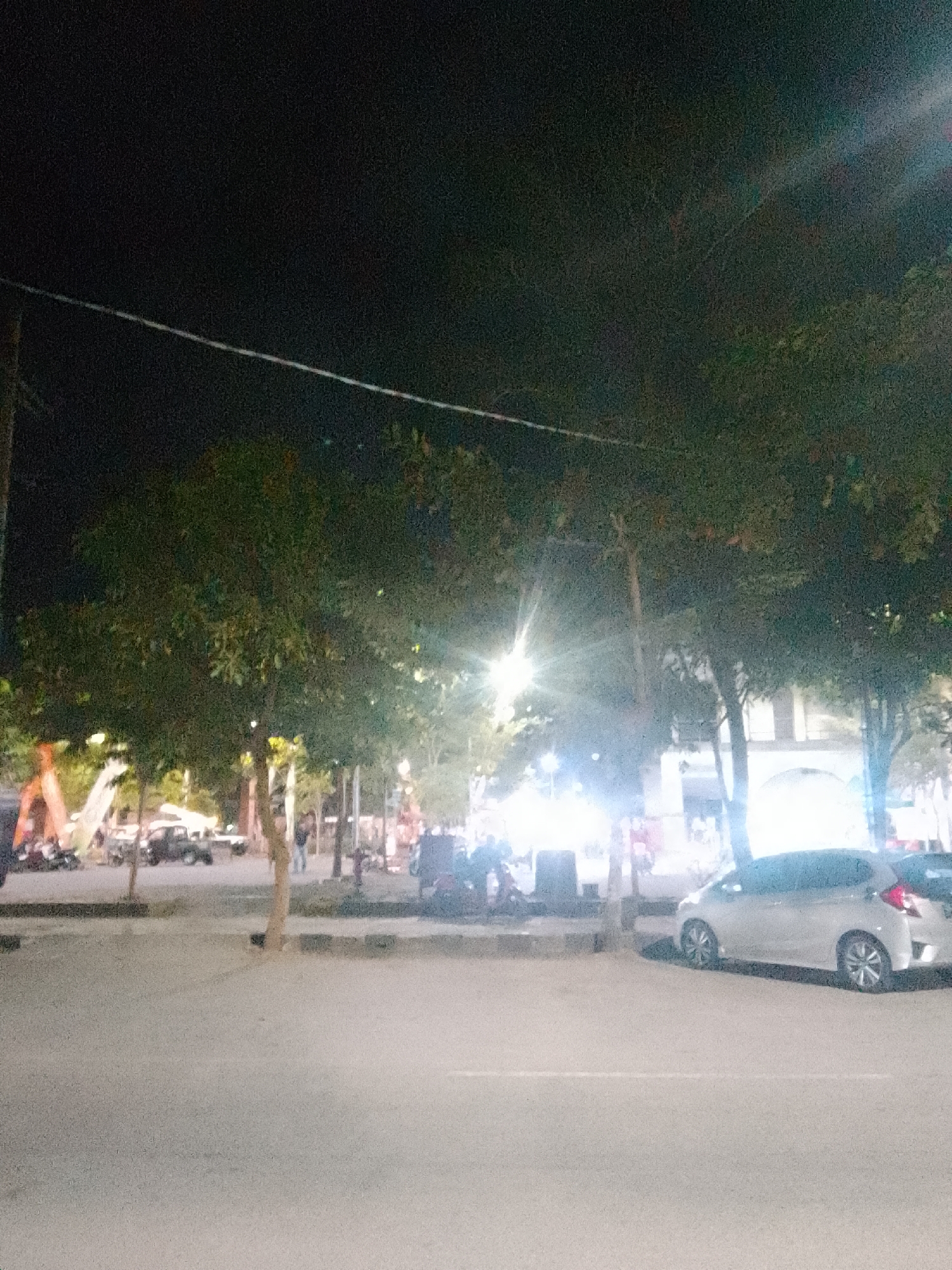 This is a low-light photograph taken at night, possibly in the city of Trenggalek, Indonesia. The scene depicts a street or plaza area with several trees lining the side of a road. In the foreground, there is a paved surface, likely a street or sidewalk, with a light-colored car parked on the right side of the image. The car appears to be a silver hatchback. Behind the trees, there are indications of buildings and streetlights, contributing to the overall illuminated but somewhat hazy atmosphere. Several people are visible in the background, some walking and others near what appears to be a motorcycle or scooter. There is also a bright light source, possibly a street lamp, in the middle-left of the frame, creating a lens flare effect. The general impression is of a busy public space at night, with a mix of natural elements (trees) and urban infrastructure.