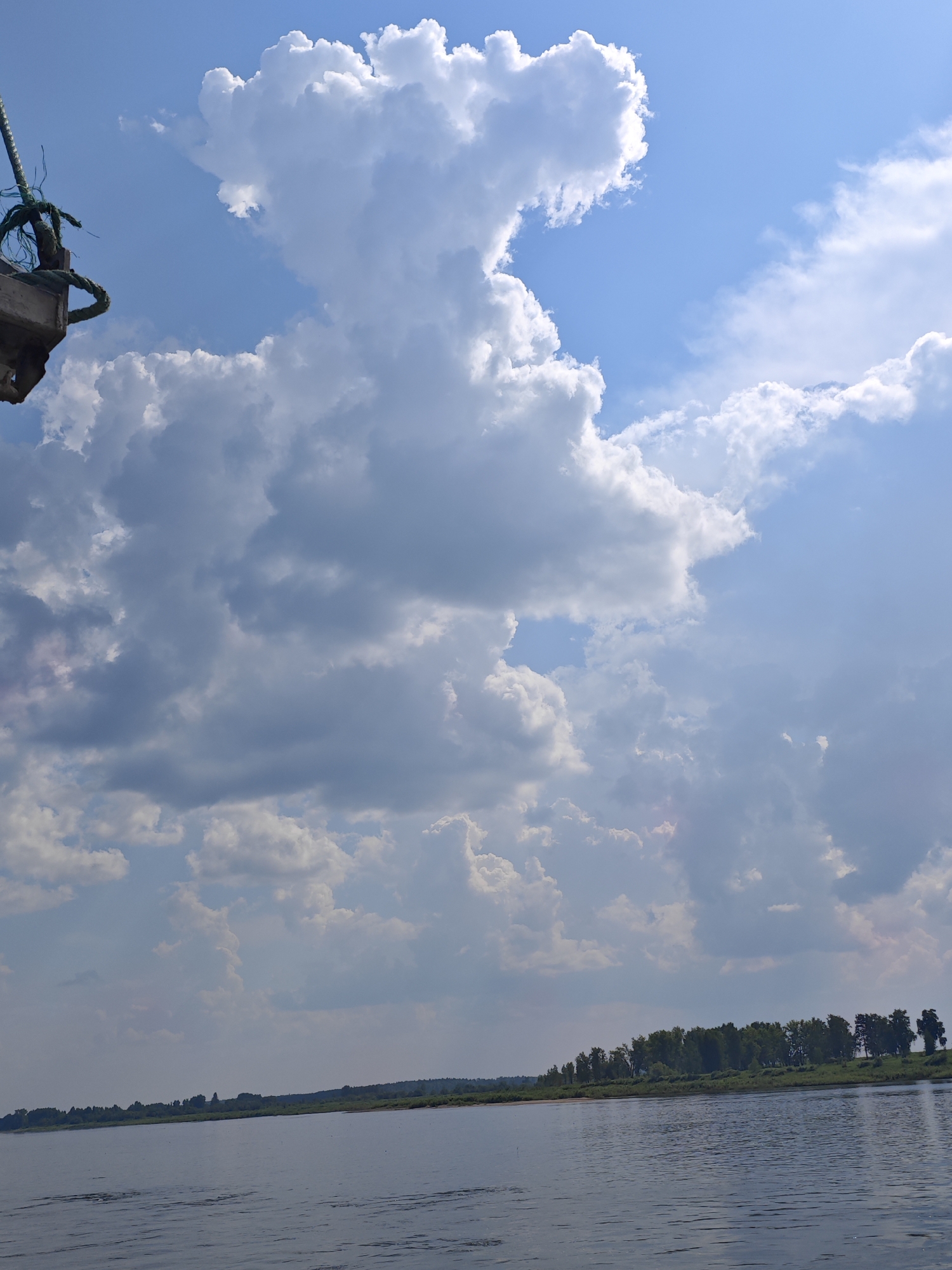 The image captures a wide, calm river in Ust'-Kem', Russia, under a sky filled with dramatic cumulus clouds. The water is a deep blue-gray, reflecting the sky and showing slight ripples on its surface. The far bank of the river is lined with a dense green forest, with a narrow strip of sand visible in places. The sky is a vibrant blue, with large, fluffy white clouds dominating the upper portion of the frame. To the left, a portion of what appears to be a boat or dock structure is visible, with a thick, weathered rope attached. The overall scene conveys a sense of natural beauty and tranquility.