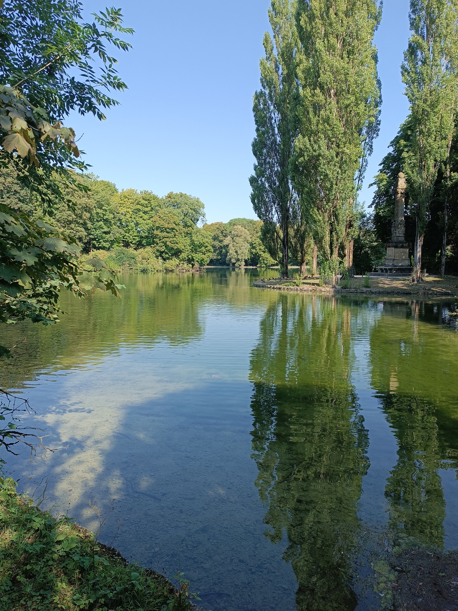 The image depicts a serene lake with clear, shallow water near the shore, revealing the gravelly bottom. The lake is surrounded by lush green trees, with the foliage of some branches reaching over the water in the foreground. Tall, slender trees with dense green leaves dominate the right side of the frame, casting reflections on the rippling surface of the lake. A stone monument or pillar, partially obscured by trees, stands on a small island or peninsula in the middle distance. The sky is a clear, bright blue. The scene appears to be a park or garden setting. This is likely the Westpark in Munich, Germany, given the presence of tall trees, a lake, and a monument in a park environment. No people are visible in the image.