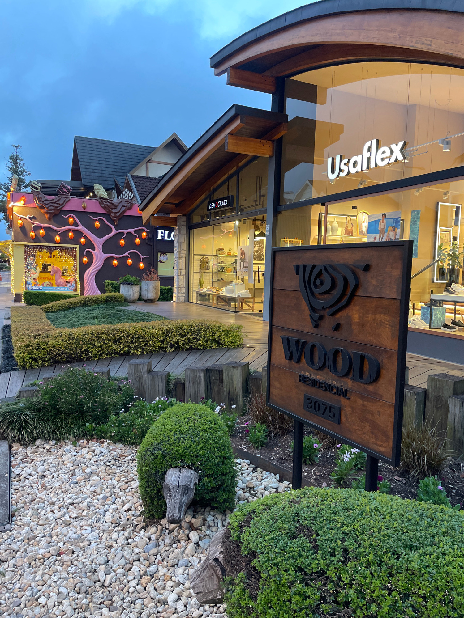The image depicts an exterior view of a shopping area in Gramado, Brazil, at dusk. The architecture features natural wood and stone elements with a modern, curved roofline. A prominent storefront displays the "Usaflex" logo. Adjacent to it, another shop has an artistic facade with a decorative tree illuminated by orange lights. A wooden sign with a stylized logo reads "WOOD RESIDENCIAL 3075". The foreground is landscaped with a variety of plants, including manicured hedges, gravel paths, and decorative wooden posts. The sky is overcast and the ambient light suggests the transition from day to night, with interior lights of the shops illuminating the scene. There are no people visible in the frame.