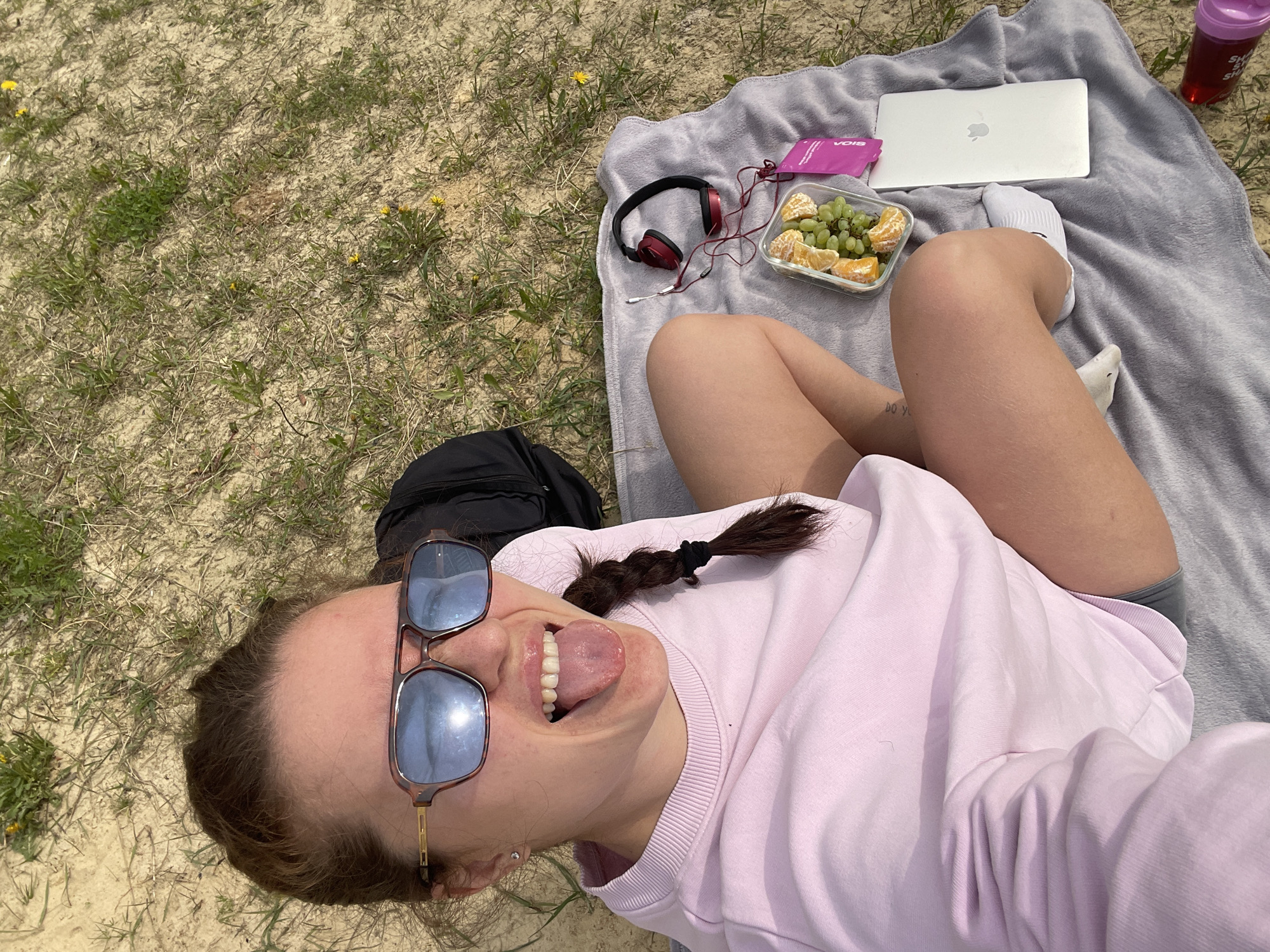 A young woman is lying on a gray blanket on a grassy surface, possibly in Misaylovo, Russia. She is wearing a light pink sweater, gray shorts, and white socks. She is sticking her tongue out and wearing sunglasses with a brown frame and blue lenses. She has brown hair pulled back in a braid. Beside her on the blanket is a laptop, a container of oranges and grapes, headphones, and a pink packet. A black backpack is positioned behind her head. A pink water bottle sits near the laptop. The ground is sandy with patches of grass and small yellow flowers.
