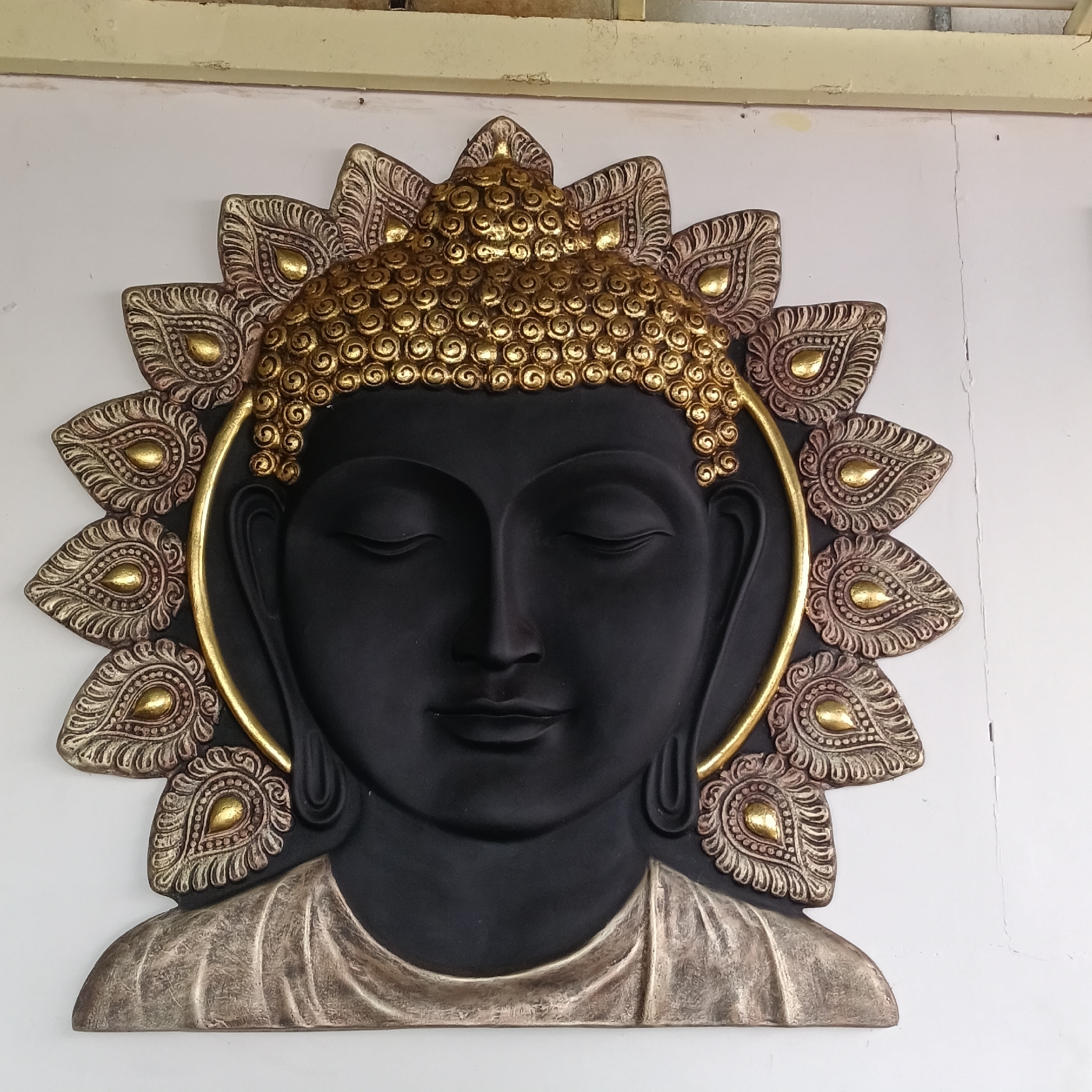 A wall-mounted Buddha statue is depicted. The face is black with closed eyes. The top of the head has a golden coil design. The statue is surrounded by a golden halo and ornate lotus petals. The wall appears to be white. Based on the location of the image in Mandrem, India, this could be a religious decoration in a home, business, or temple.