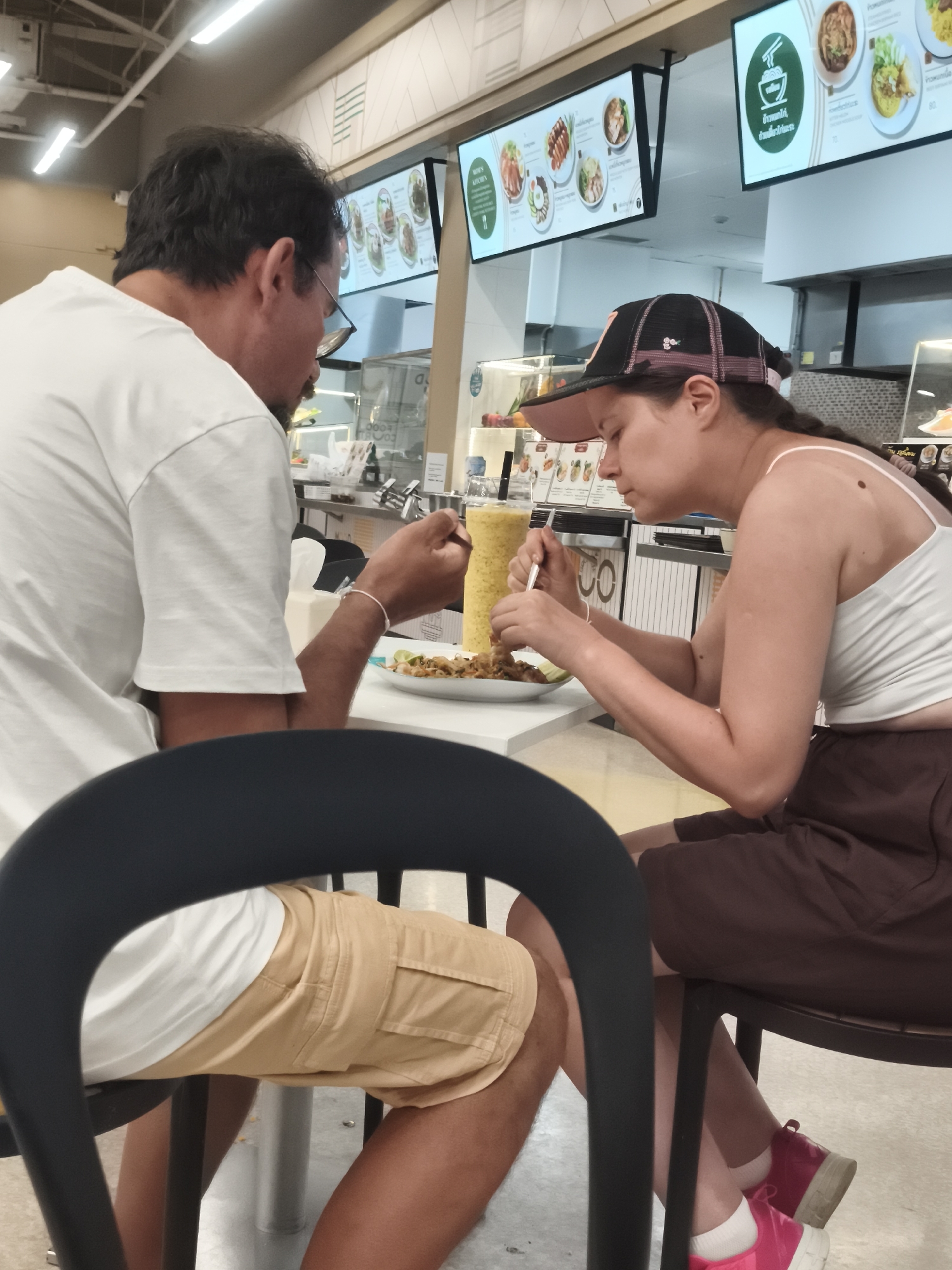 In Baan Lamai, Thailand, two individuals are seated at a white table, engaged in eating. The person on the left, a man with dark hair and glasses, wears a white t-shirt and is seen holding a fork. To his right, a woman wearing a black baseball cap with pink accents and a white tank top is also eating, using a fork and spoon. In front of them are plates of food and a tall, yellow beverage in a plastic cup with a straw. The environment appears to be a food court or restaurant, with digital menus displaying food images and prices hanging from the ceiling. The lighting suggests an indoor setting during the daytime.