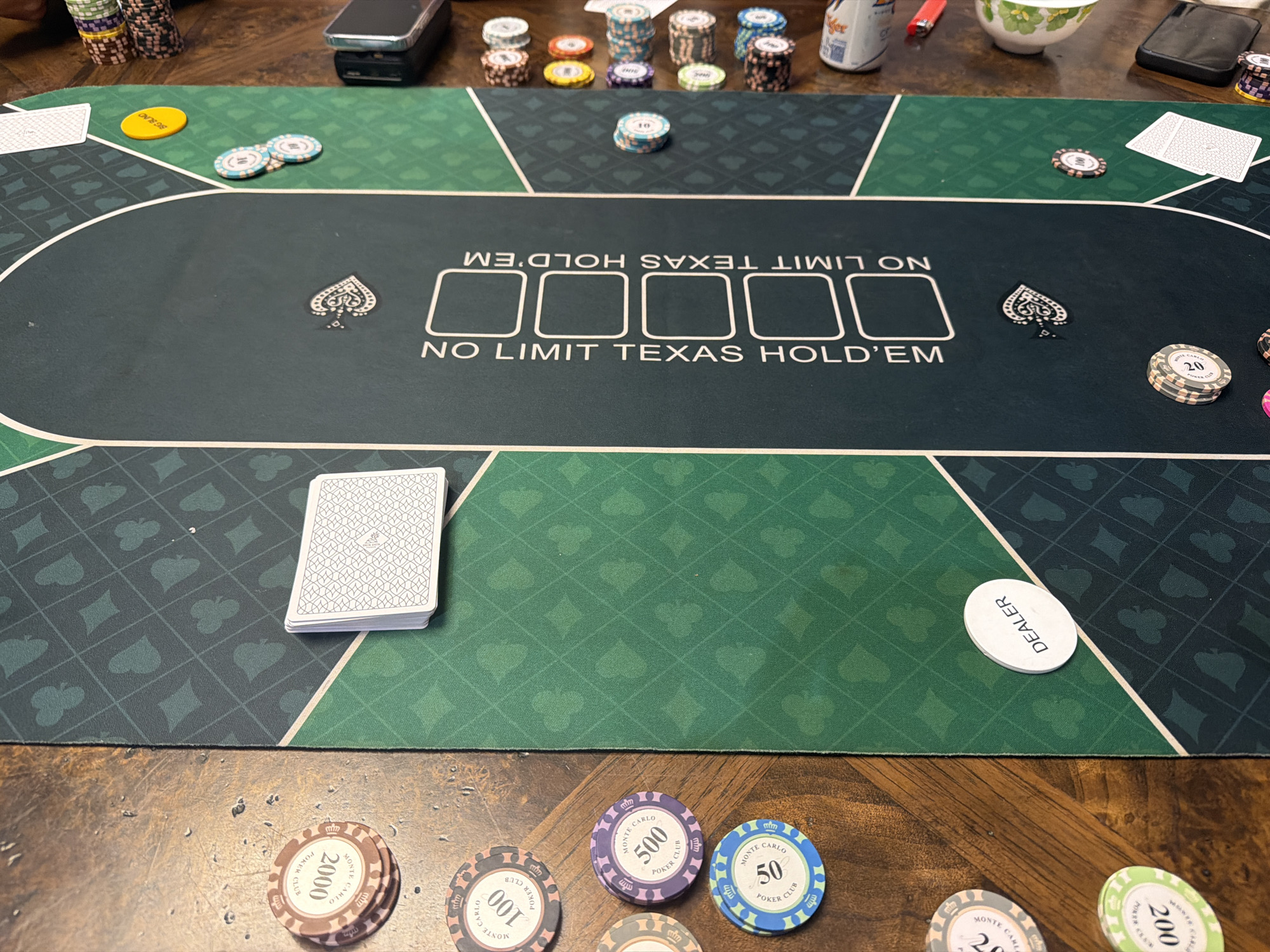 The image depicts a close-up, high-angle view of a poker table setup. The table surface is a dark green mat with a repeating pattern of diamond and spade card suits. Across the center of the mat, the words "NO LIMIT TEXAS HOLD'EM" are prominently displayed in white lettering, with five empty rectangular slots above them, likely for community cards.
In the foreground, on a wooden table, are several stacks of poker chips. These chips vary in color and denomination. Visible denominations include 50, 100, 200, 500, and 2000, with "MONTE CARLO POKER CLUB" inscribed on some. There is also a white disc labeled "DEALER" positioned on the green mat. A deck of cards, with its back facing upwards and displaying a repeating geometric pattern, rests on the left side of the mat.
On the upper portion of the green mat, near the edge, a single yellow poker chip is visible, along with two light blue and white chips stacked together. Further up, on the wooden surface, are stacks of various colored chips, a can that appears to be a beverage container, a red lighter, a small white bowl, and a black electronic device. The overall scene suggests a game of Texas Hold'em is either in progress or about to begin. There are no people visible in the image.
The location of Xuân Trường - Đà Lạt, Vietnam, is not visually represented or inferable from the provided image.