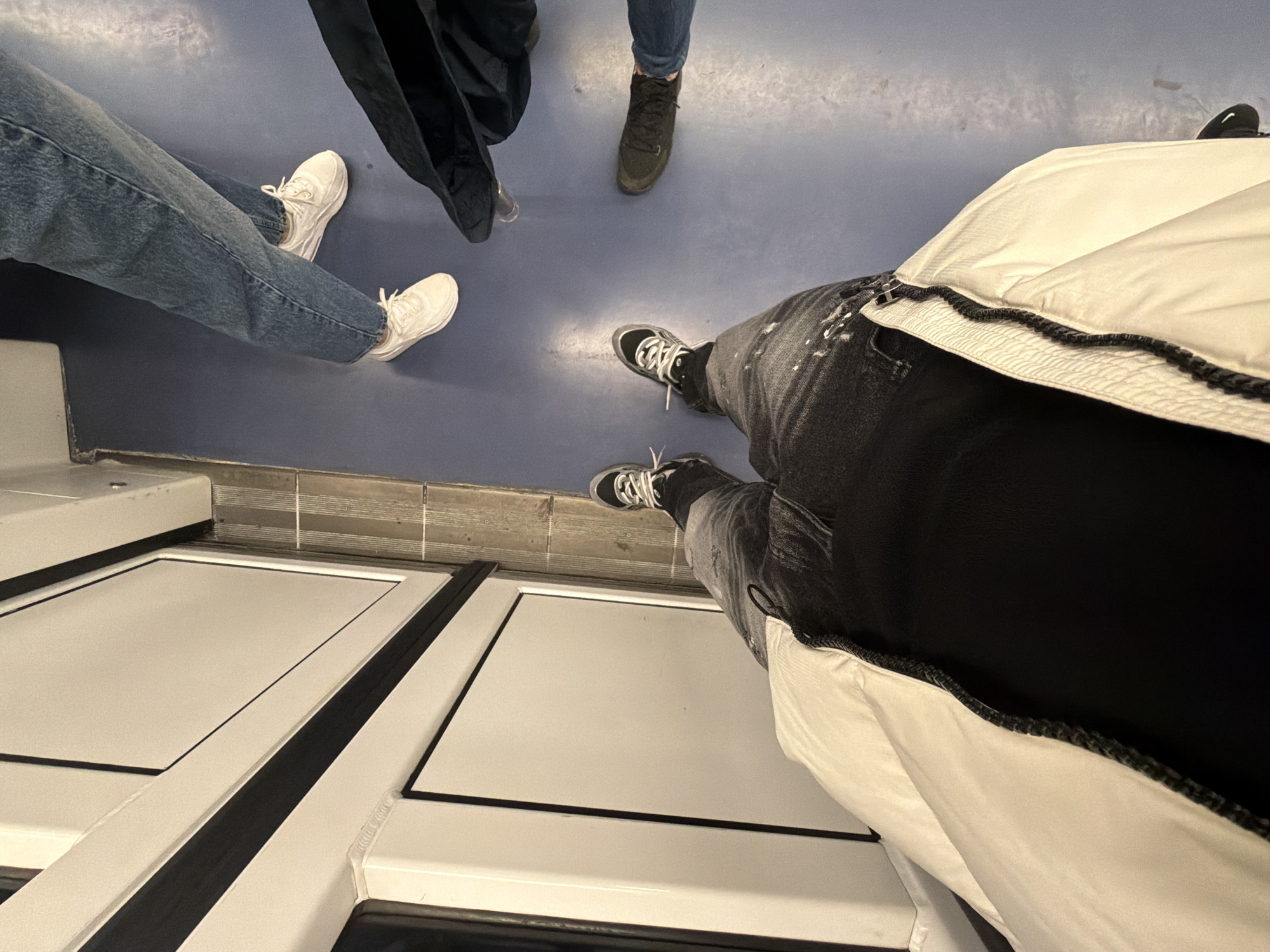 The image shows a view from above of a train car in Milan, Italy. Three people are standing on the blue floor. One person is wearing blue jeans and white sneakers. Another person is wearing dark jeans and dark-colored boots. A third person is wearing black jeans and black and white sneakers, and a white jacket. The doors of the train car are visible in the foreground. The train car is clean and well-maintained.