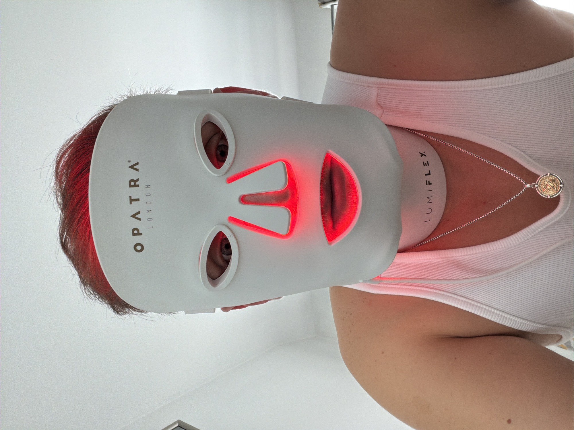 A person wears a white face mask illuminated with red light, obscuring most of their face except for the eyes. The mask has the words "OPATRA LONDON" printed on its side. The person also wears a white tank top and a necklace with a circular pendant. A white device with the word "LUMIFLEX" printed on it rests on their neck beneath the mask. The background appears to be a plain white wall. The location is not ascertainable from this image.